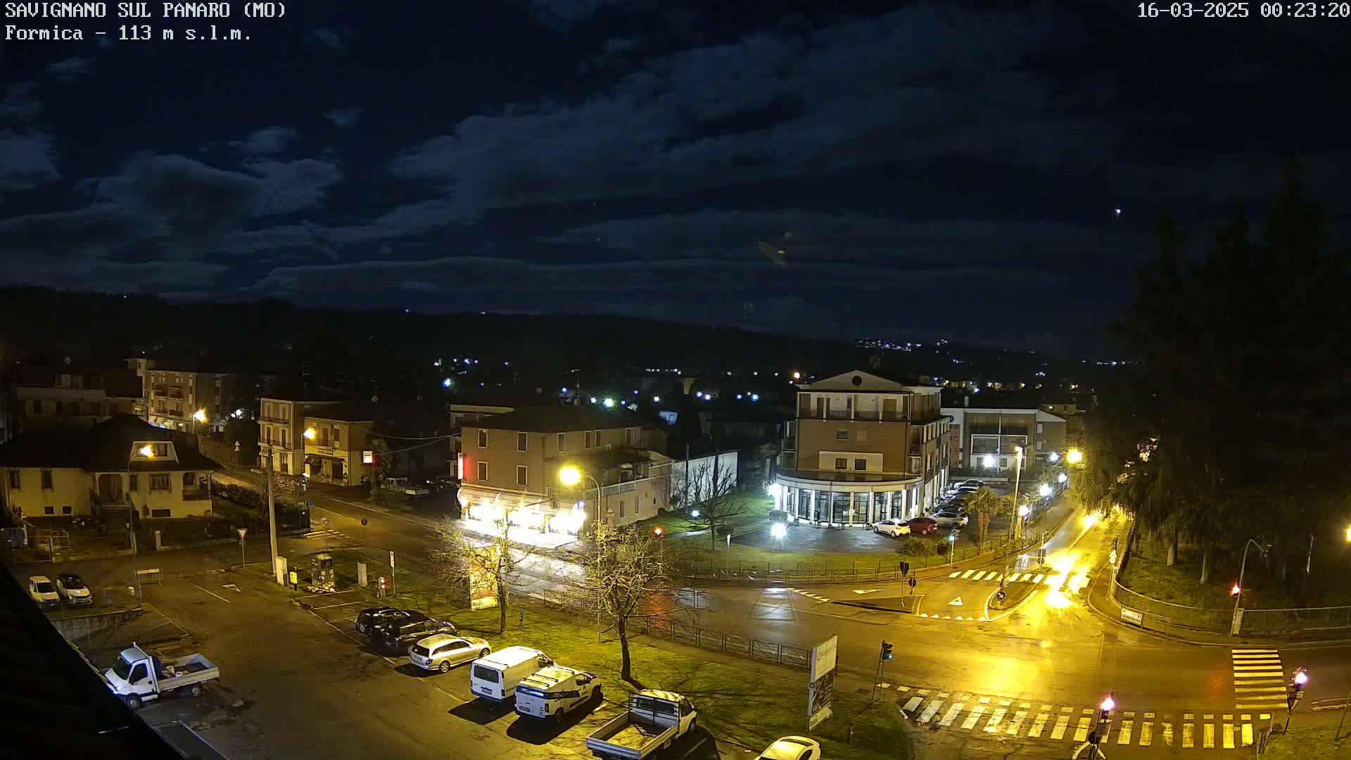 A nighttime aerial view of a town square with parked cars and streetlights under a partly cloudy sky.