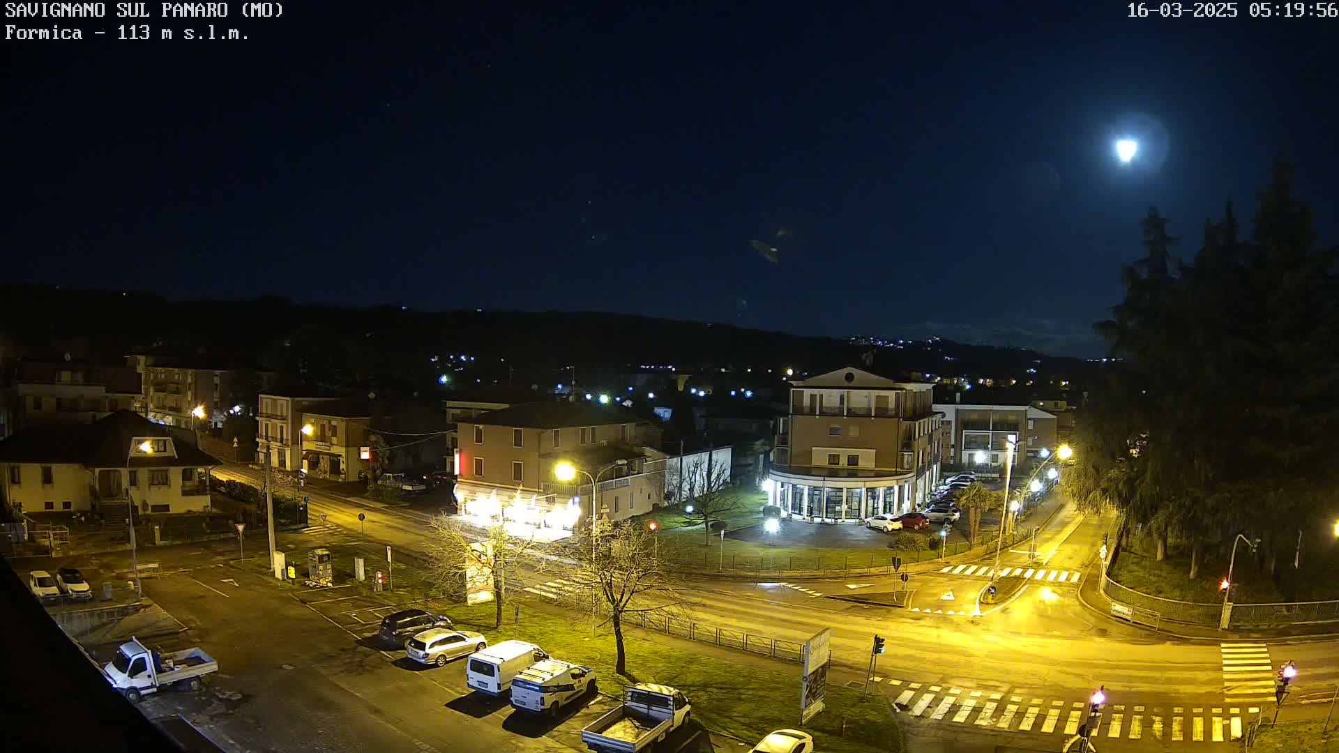 A nighttime, overhead view of a town square with parked cars and illuminated streets under a bright moon.