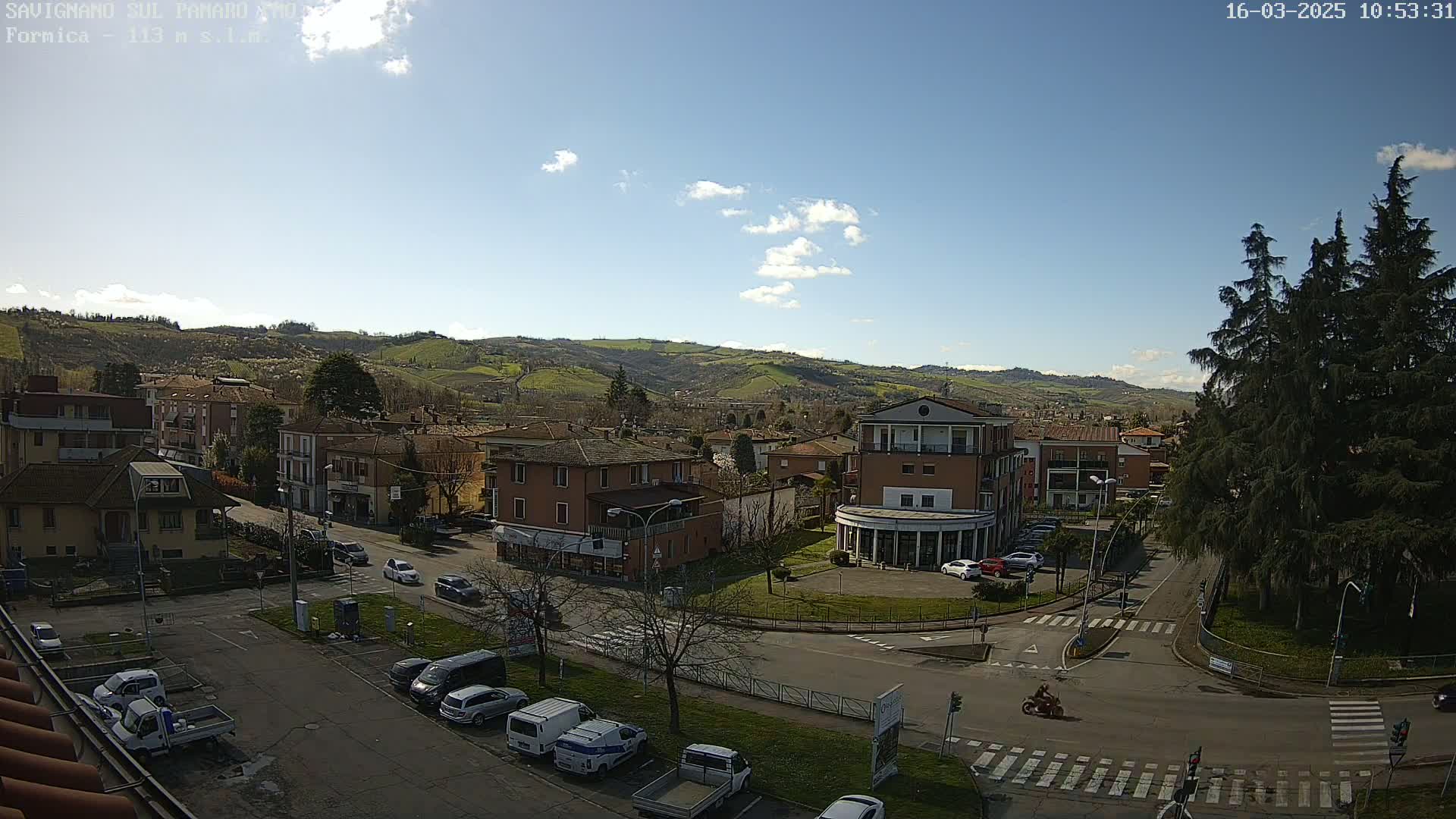 A sunny day reveals a view of a town nestled in rolling hills, with cars parked in a lot and a motorcyclist driving on a street below.