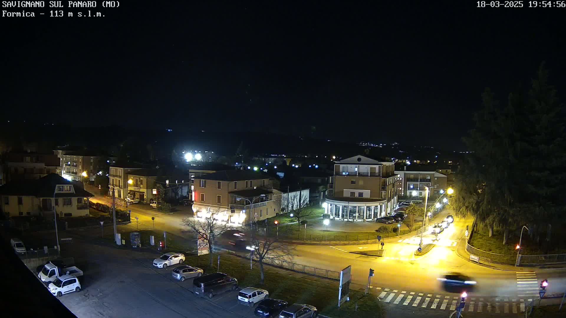 A nighttime, high-angle view of a town square with several cars parked and driving, illuminated by streetlights under a clear, dark sky.