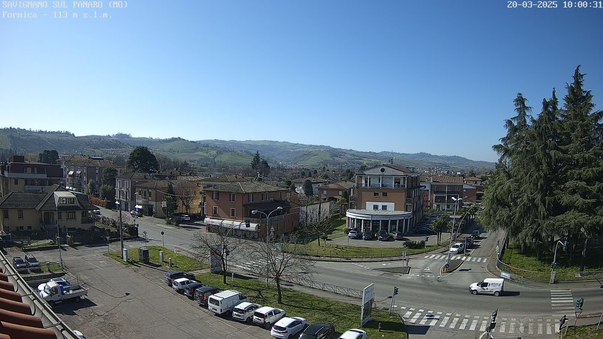A sunny day reveals a town nestled in a valley, surrounded by rolling hills and featuring a street intersection with parked cars and a few buildings.