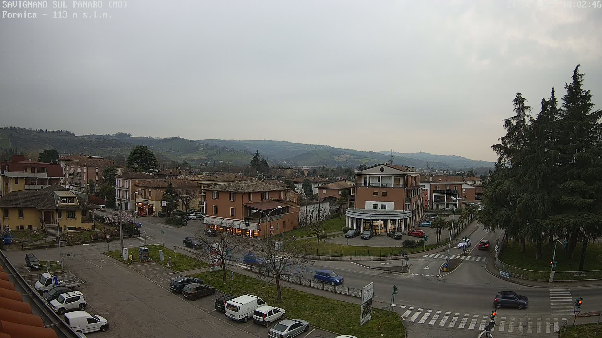 An overcast day reveals a view of a town square with parked cars and buildings, surrounded by rolling hills in the distance.