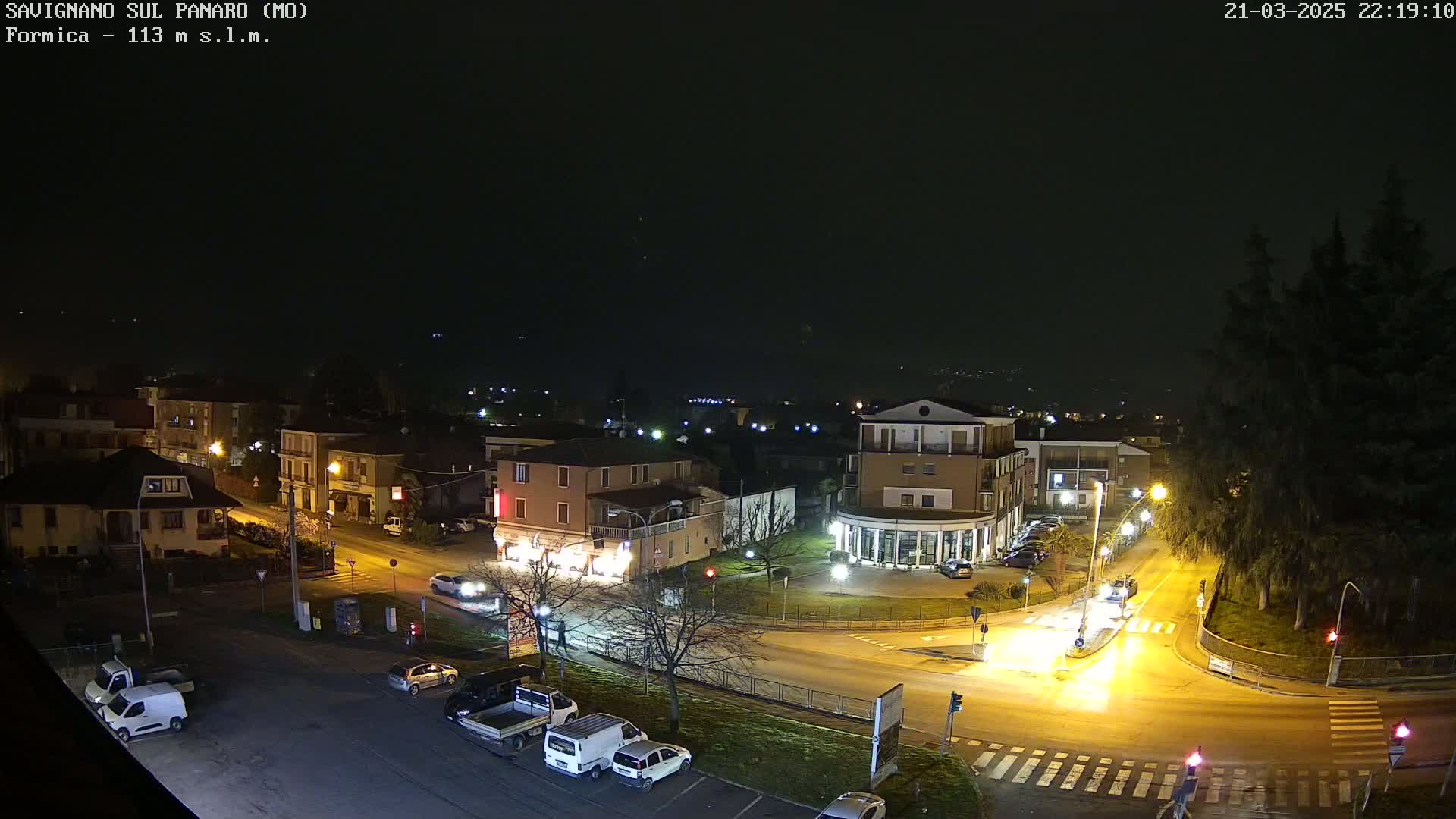 A nighttime aerial view of a town square with several cars parked and driving on the roads, under a dark, clear sky.