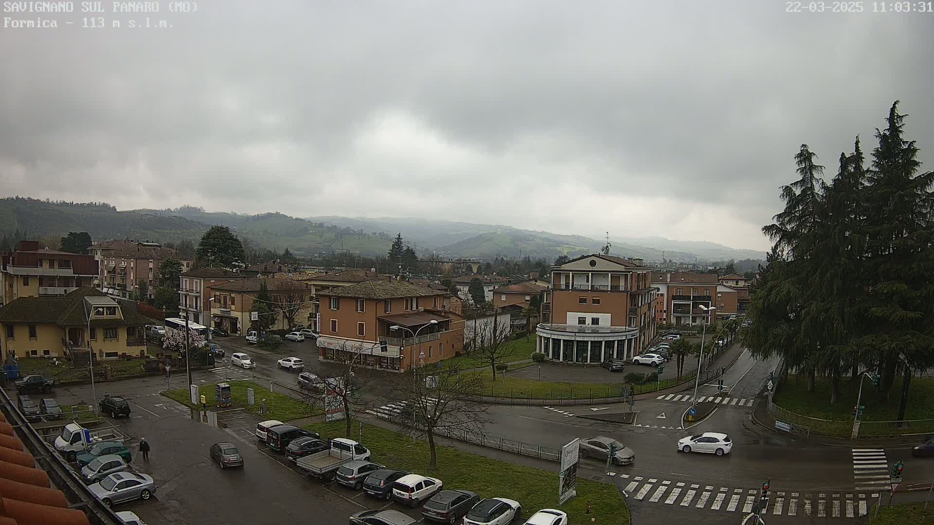 A view of a town in a valley under an overcast sky, showing buildings, cars, and trees, with wet streets indicating rain.