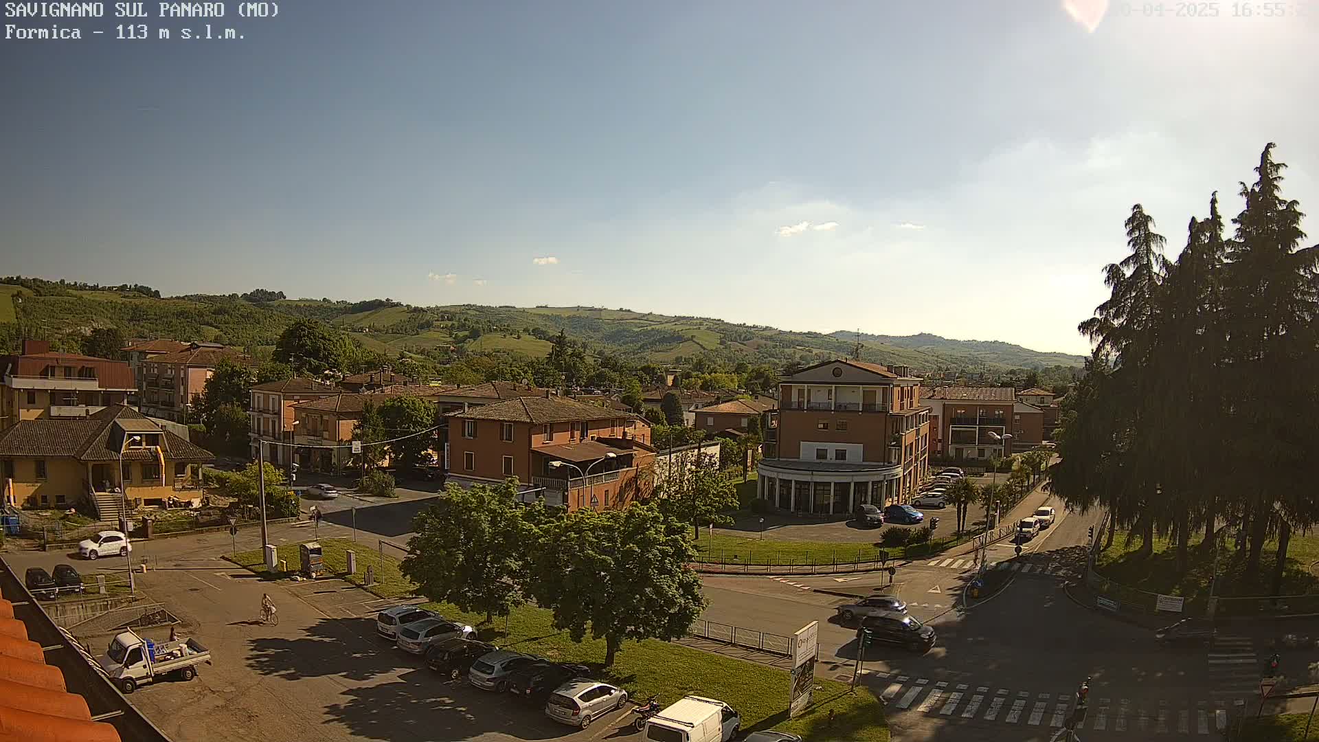 A sunny view of a town nestled in a valley, surrounded by rolling green hills and tall evergreen trees, with cars parked in a lot and driving on the roads.