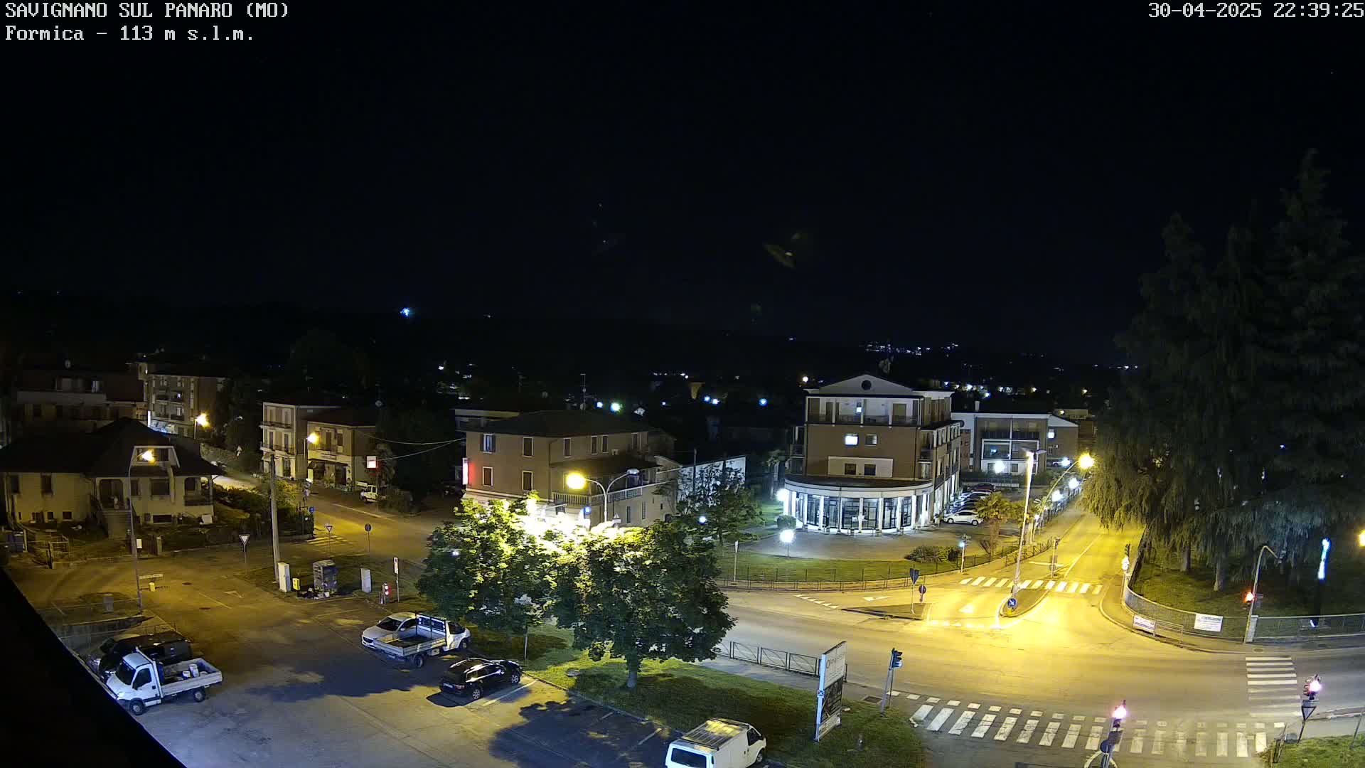 A nighttime, aerial view of a town square with several buildings, vehicles, and streetlights illuminating the scene.