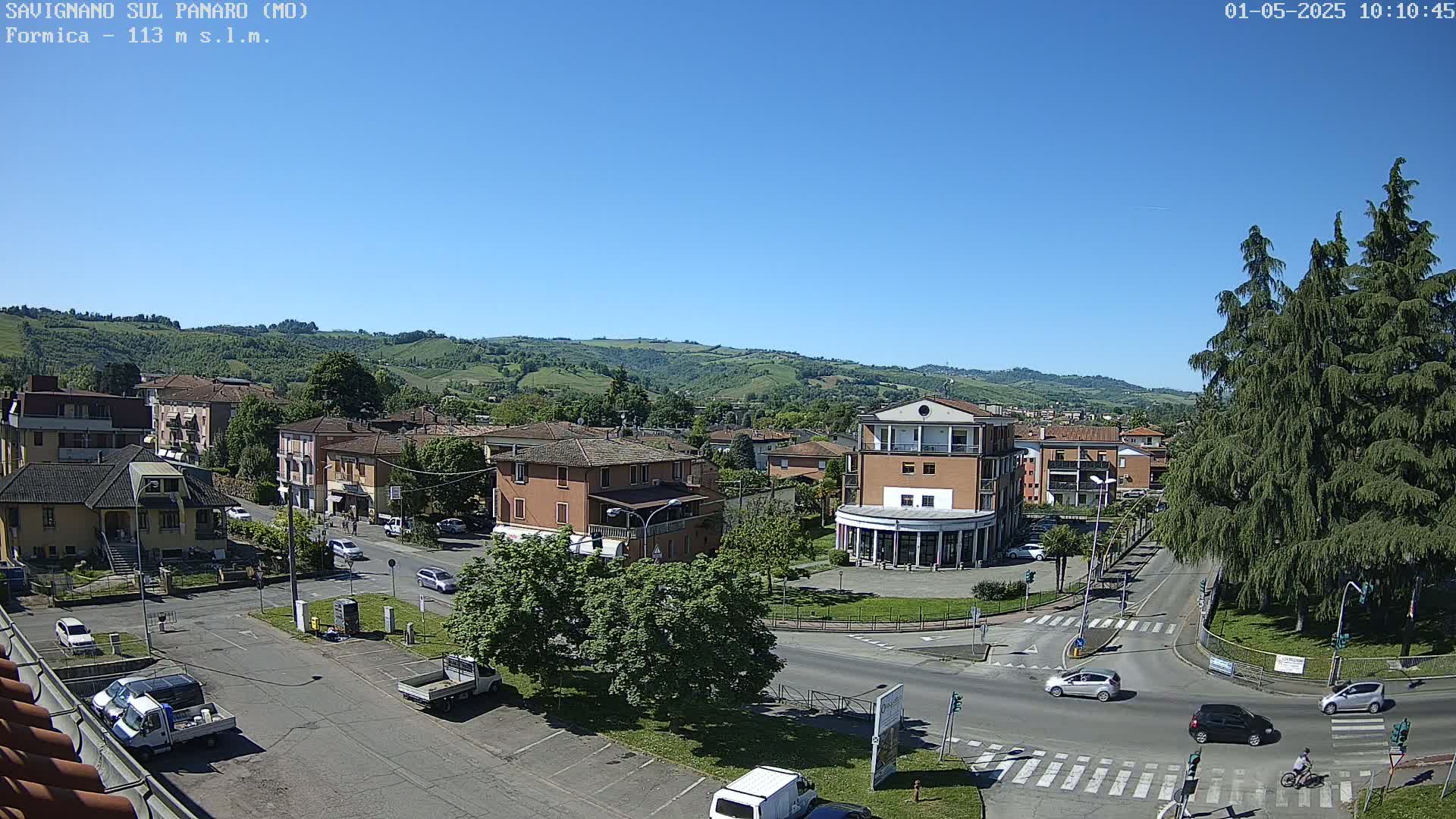 A high-angle view of a town square with buildings, cars, and trees under a clear, sunny sky, nestled in a valley surrounded by rolling hills.