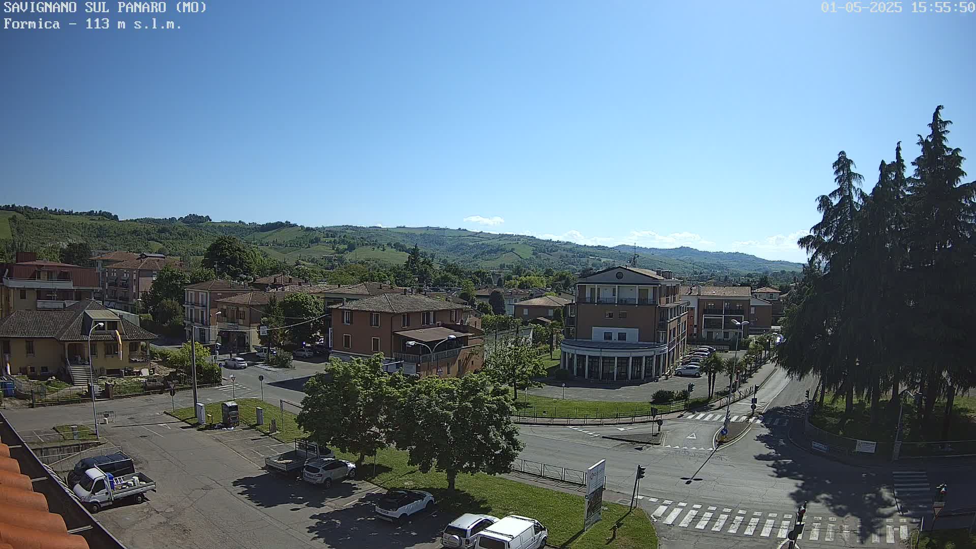 A sunny day reveals a town nestled in a valley, surrounded by rolling green hills and tall evergreen trees, with cars parked in a square and roads intersecting.