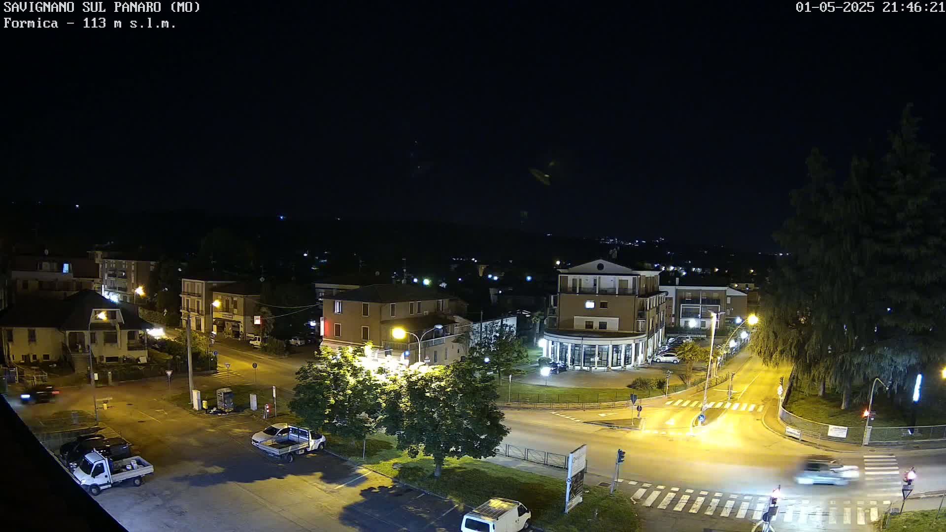 A nighttime, high-angle view of a town square with several buildings, vehicles, and streetlights illuminated under a clear, dark sky.