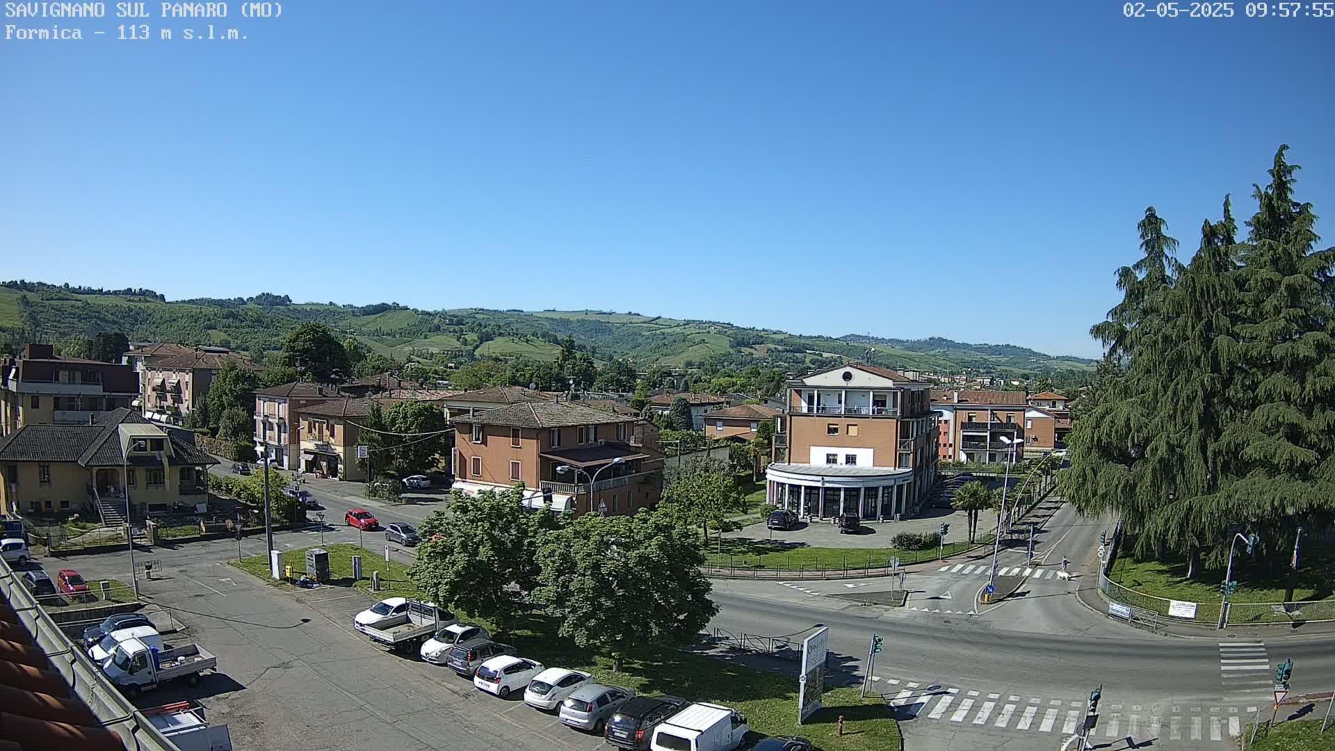 A sunny day reveals a town square surrounded by low-rise buildings and rolling green hills in the background.