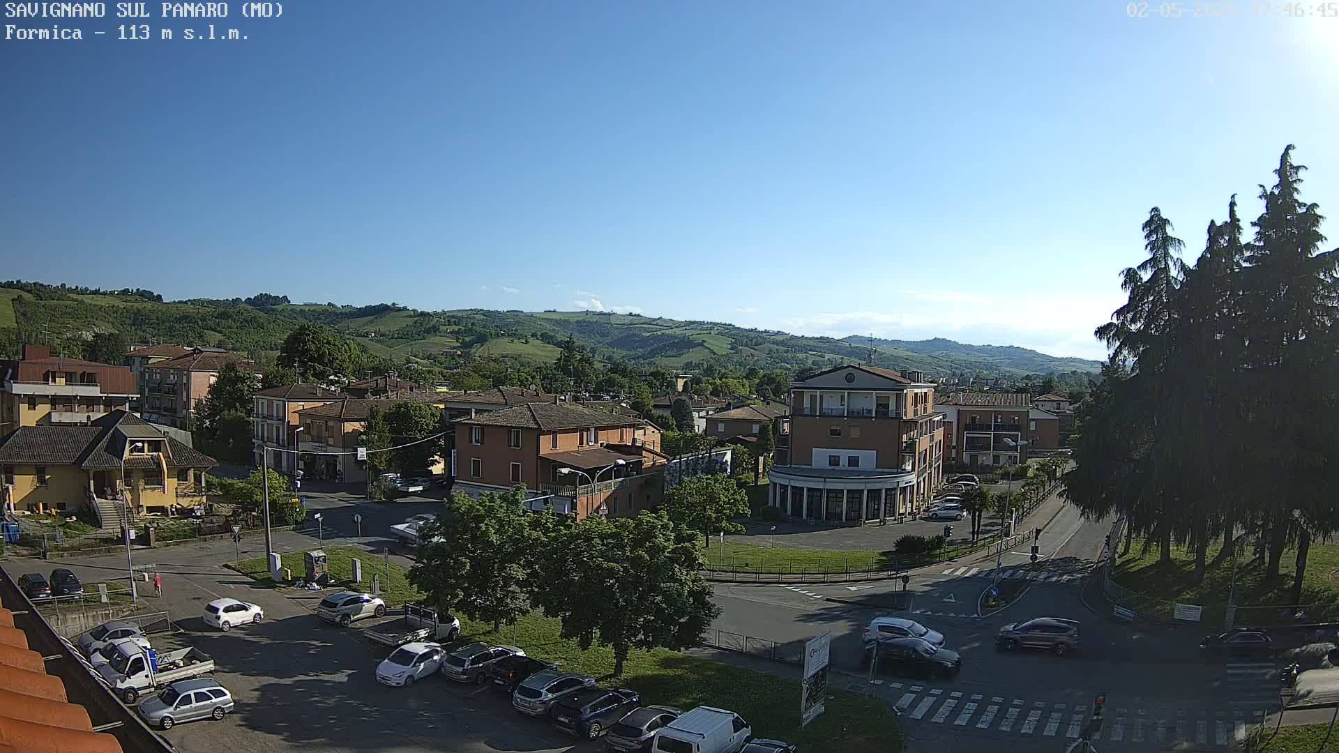 A sunny day reveals a town nestled in a valley, with cars parked along streets and several buildings visible, surrounded by lush green hills.