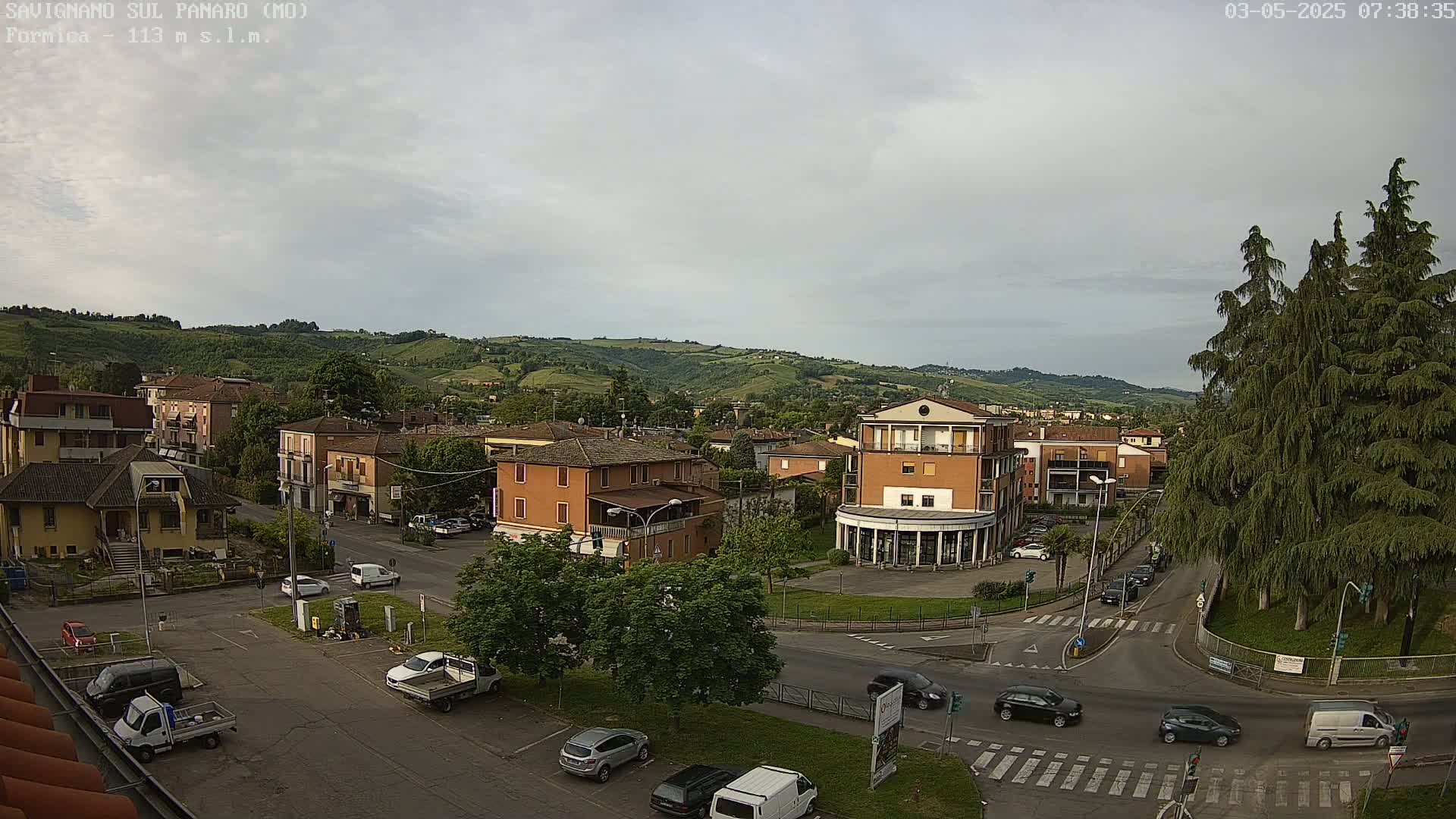 An overcast day reveals a town nestled in a valley, with cars driving along roads that intersect near a small roundabout surrounded by trees.