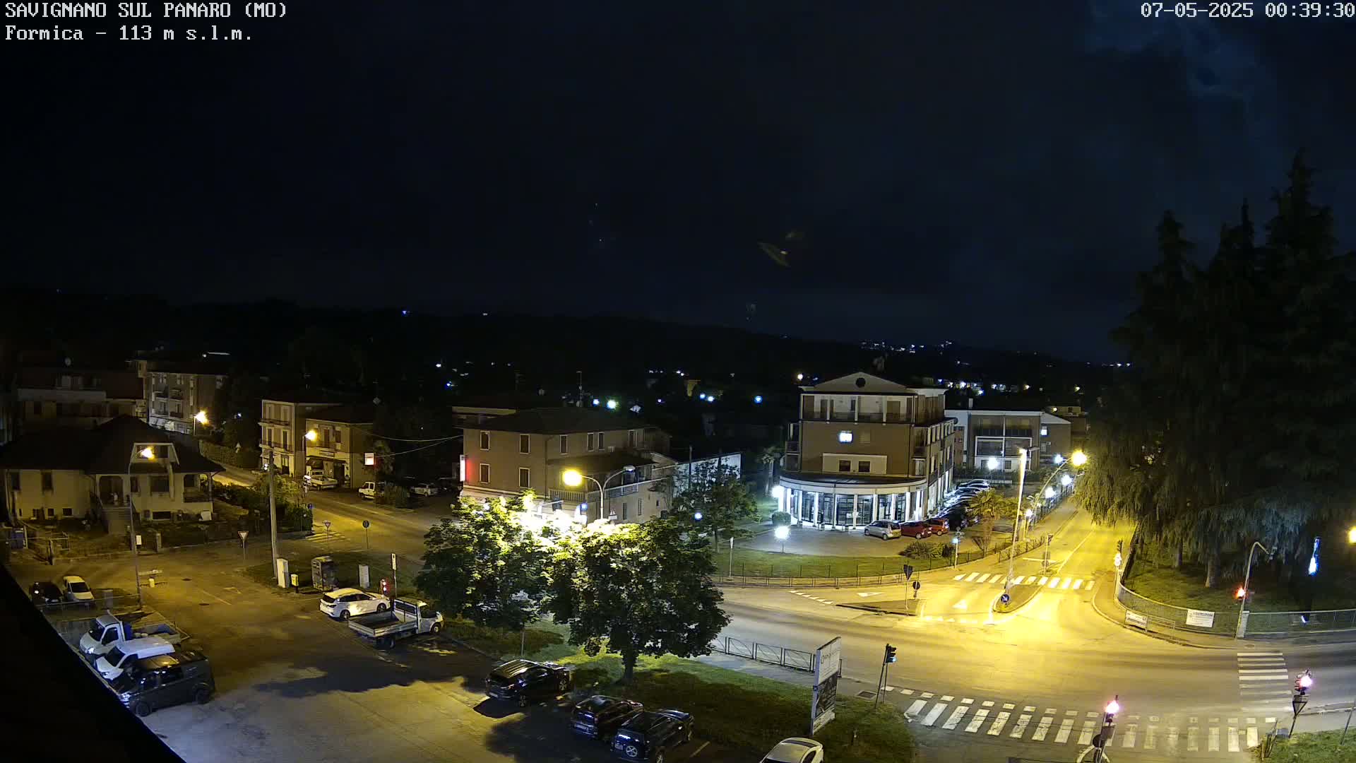 A nighttime aerial view of a town intersection with cars parked and driving, under a dark, clear sky.