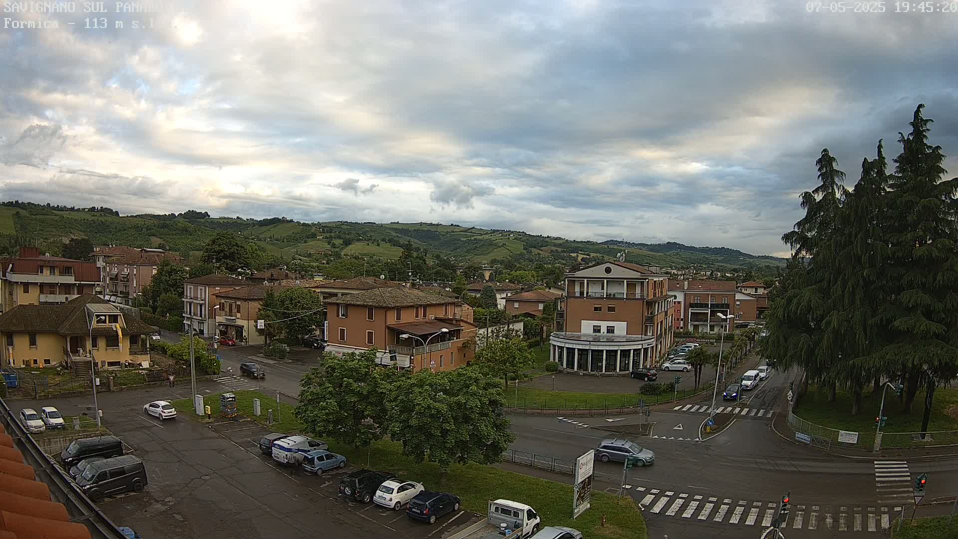 A partly cloudy daytime view of a town nestled in a valley, with cars parked and driving on roads that intersect near a small park.
