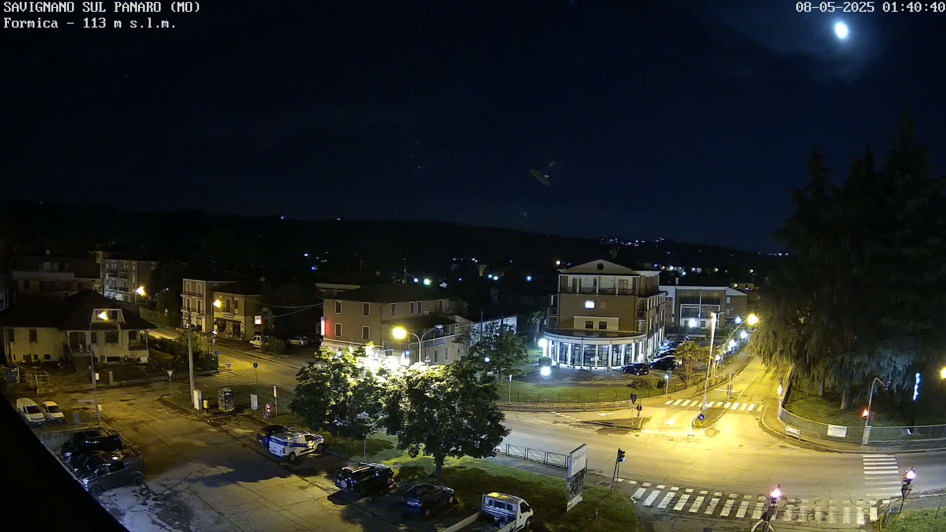 A nighttime, overhead view of a town square with cars parked along the streets under a bright moon.