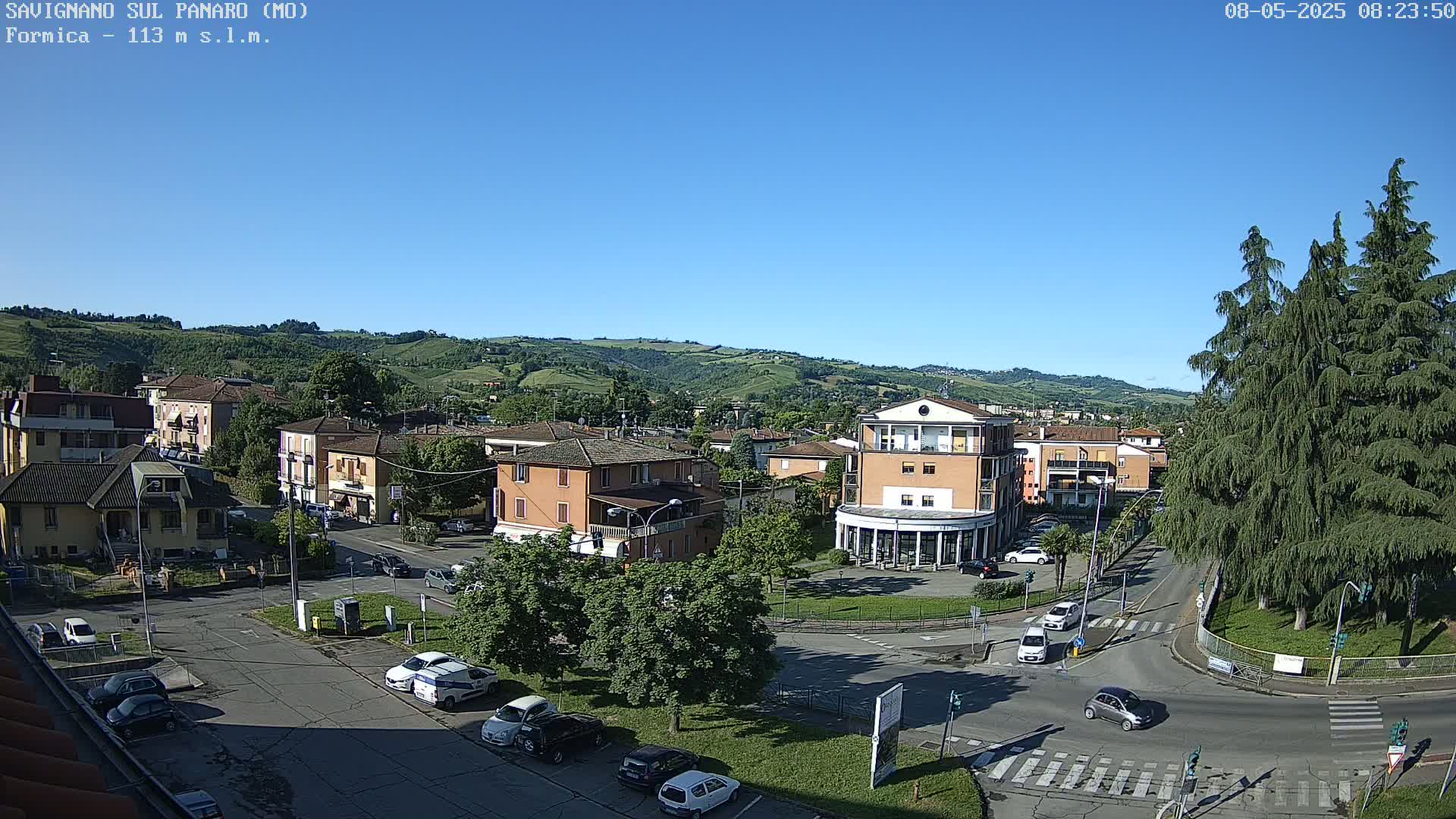 A sunny day reveals a town nestled in a valley, with cars parked along roads and buildings of varying sizes, surrounded by green hills and trees.