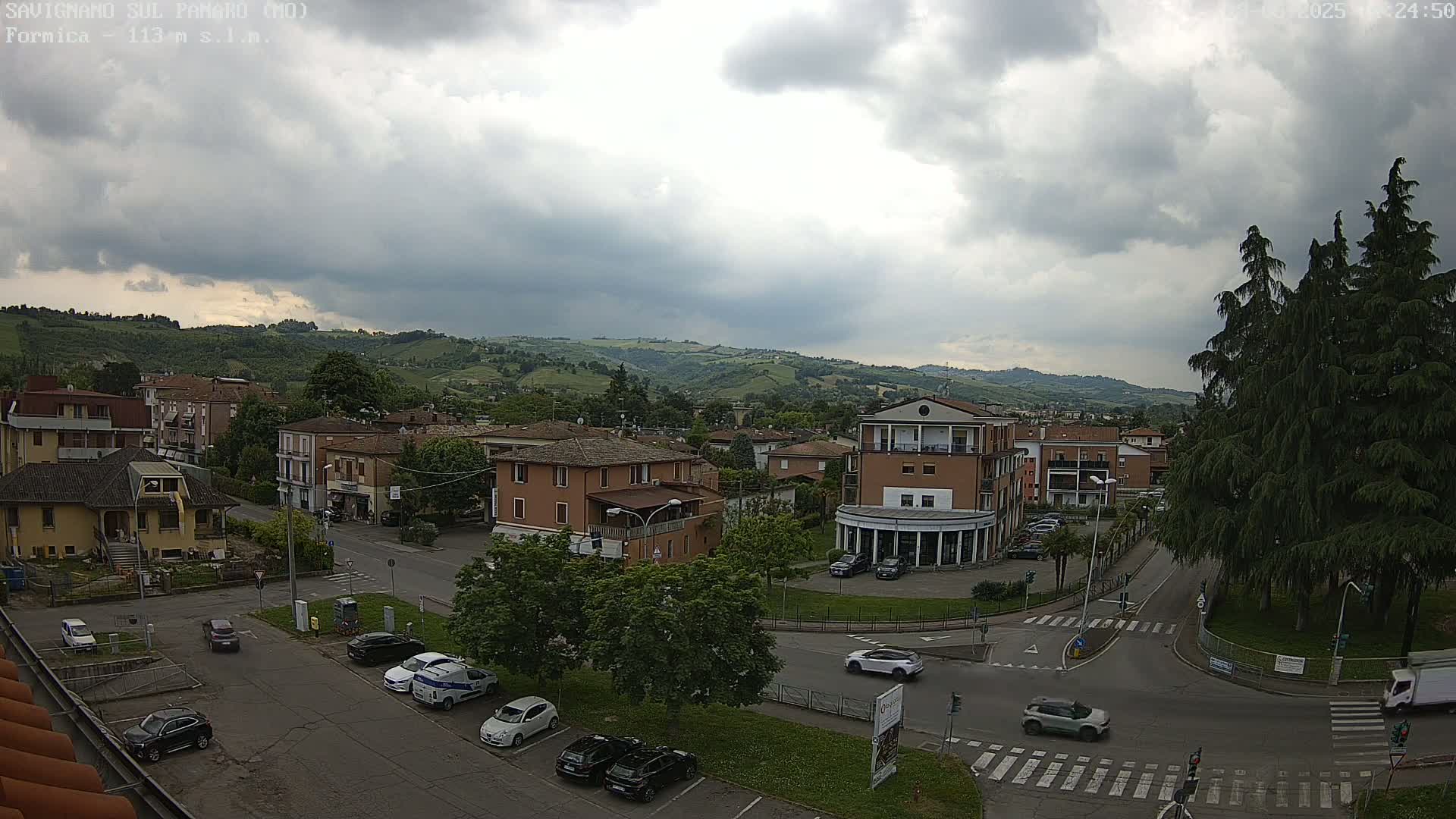 A view of a town square and surrounding buildings under a cloudy sky, with rolling hills in the background and several cars visible on the roads.