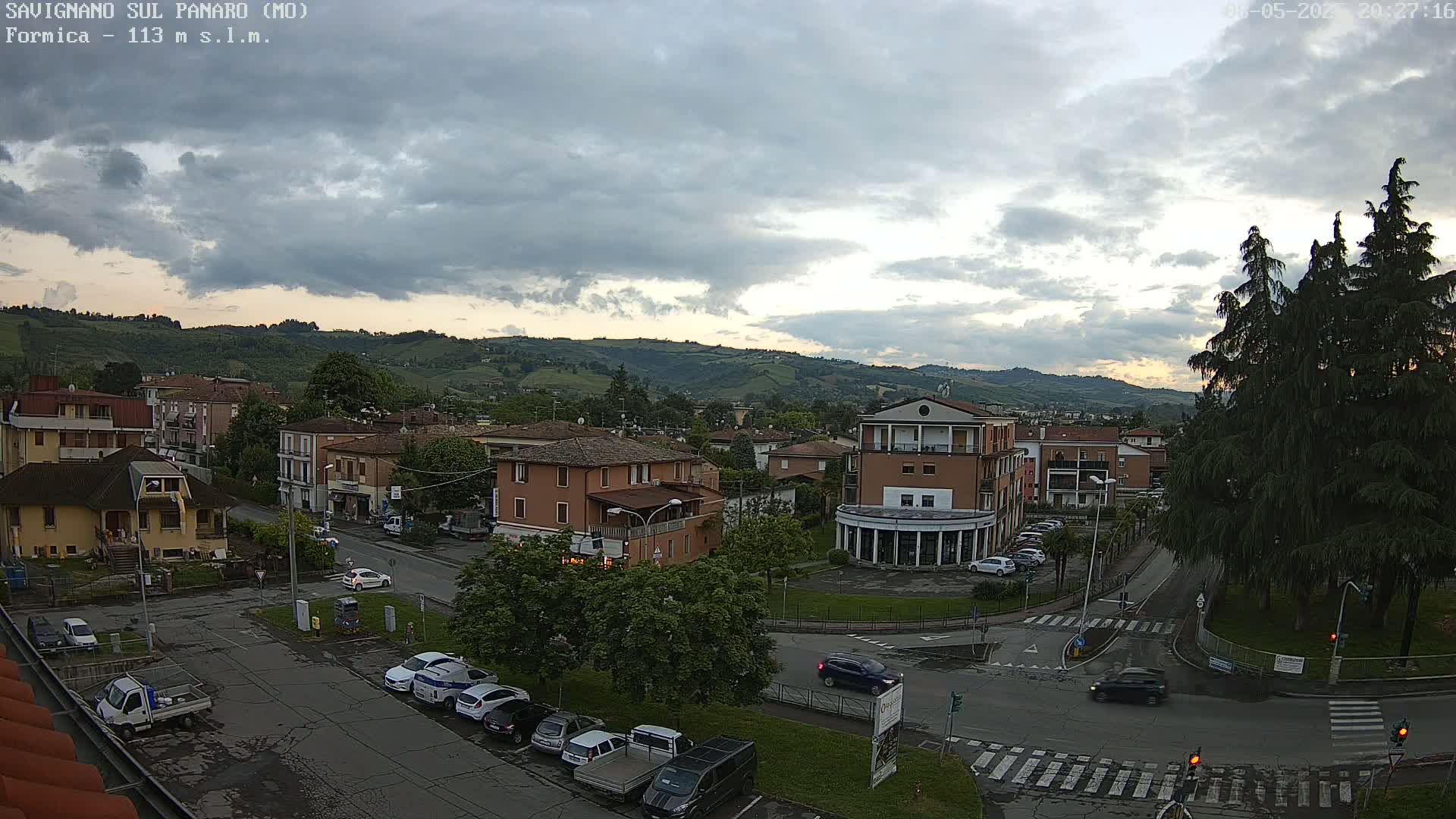 A cloudy evening view of a town nestled in a hilly landscape, with cars parked in a lot and driving on the roads below.