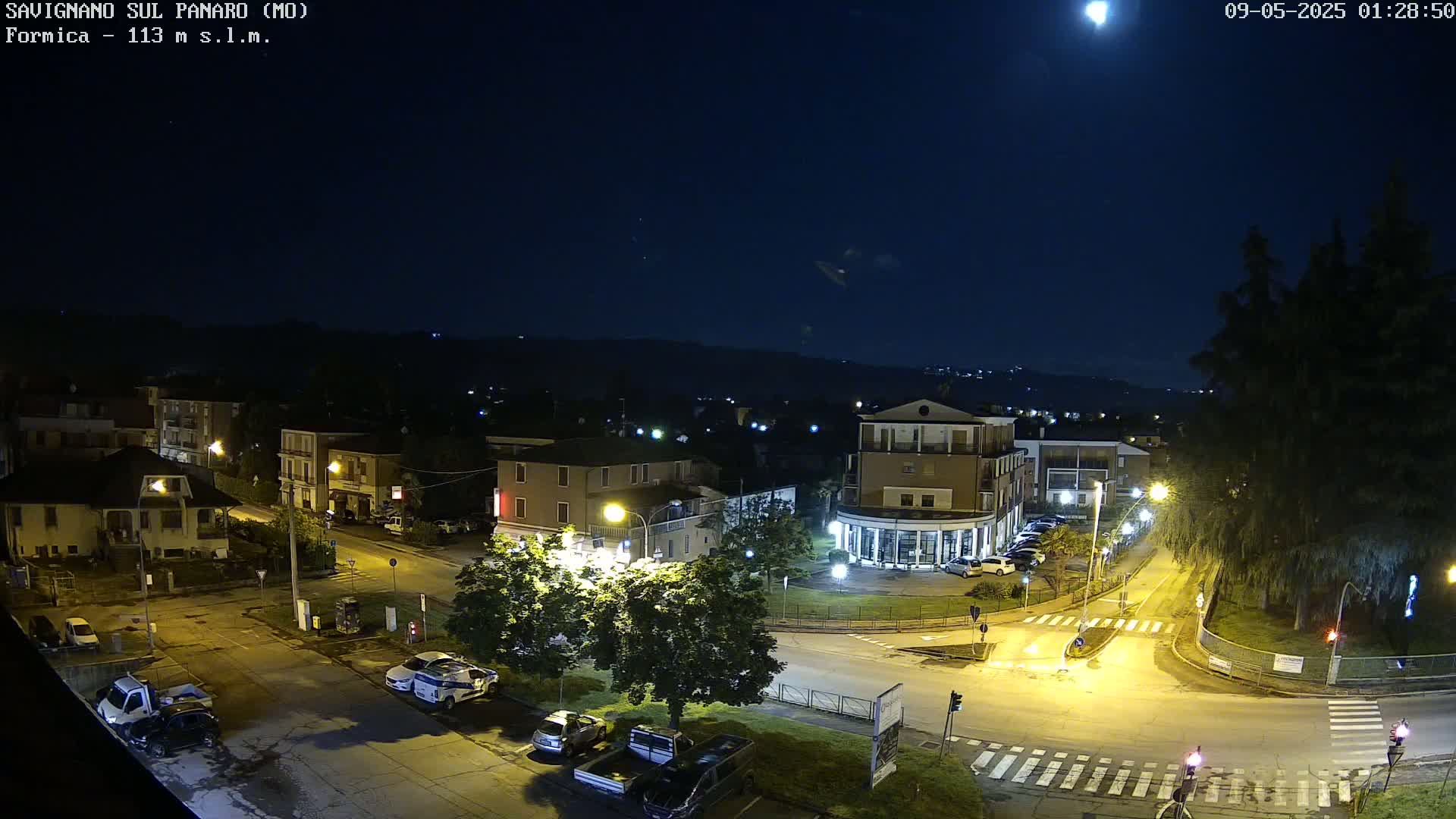 A nighttime, overhead view of a town square with several buildings, cars, and streetlights illuminated under a mostly clear sky with a bright moon.