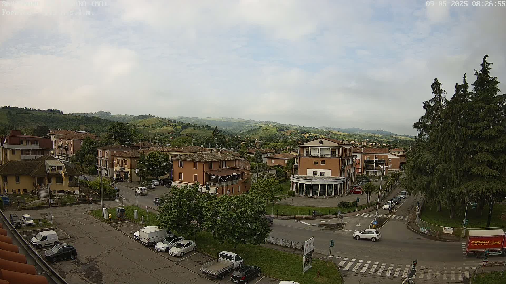 A mostly cloudy day reveals a town nestled in a valley, surrounded by green hills and featuring cars parked in lots and driving on roads that intersect near a central plaza.