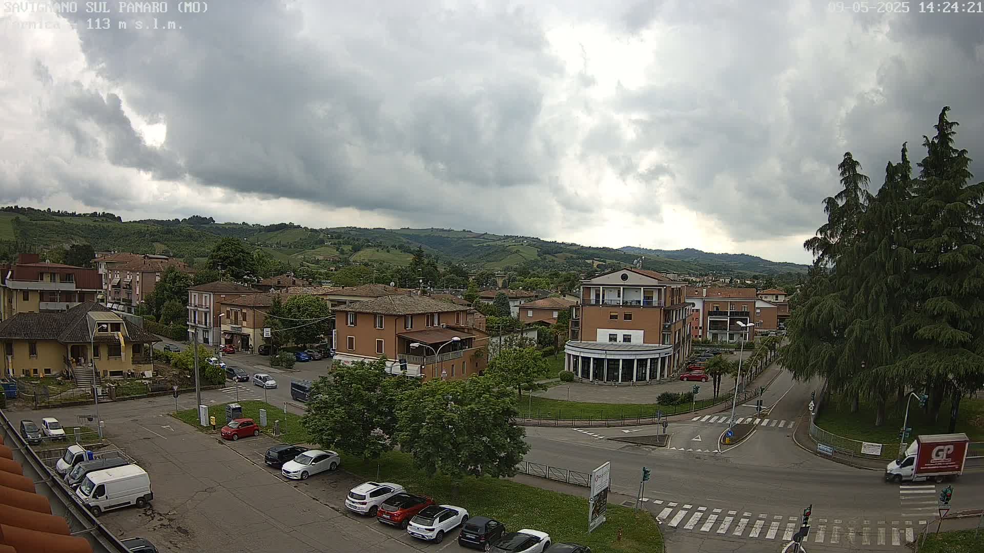 A view of a town square with overcast skies and several cars parked in the lot, nestled amongst low-lying hills.