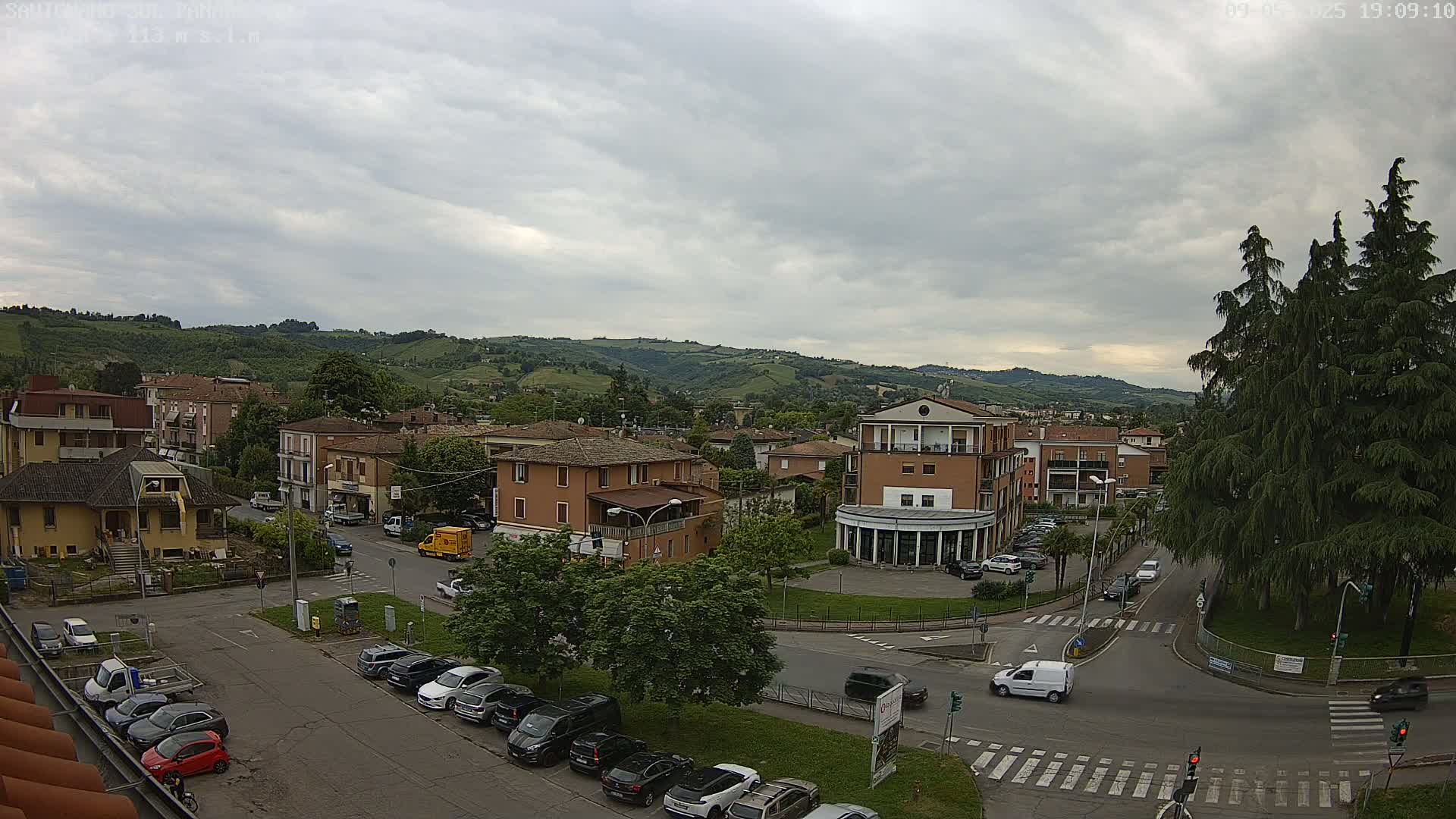 An overcast day reveals a town nestled in a valley, with cars parked along streets and a few vehicles driving through a crossroads near a large evergreen tree.