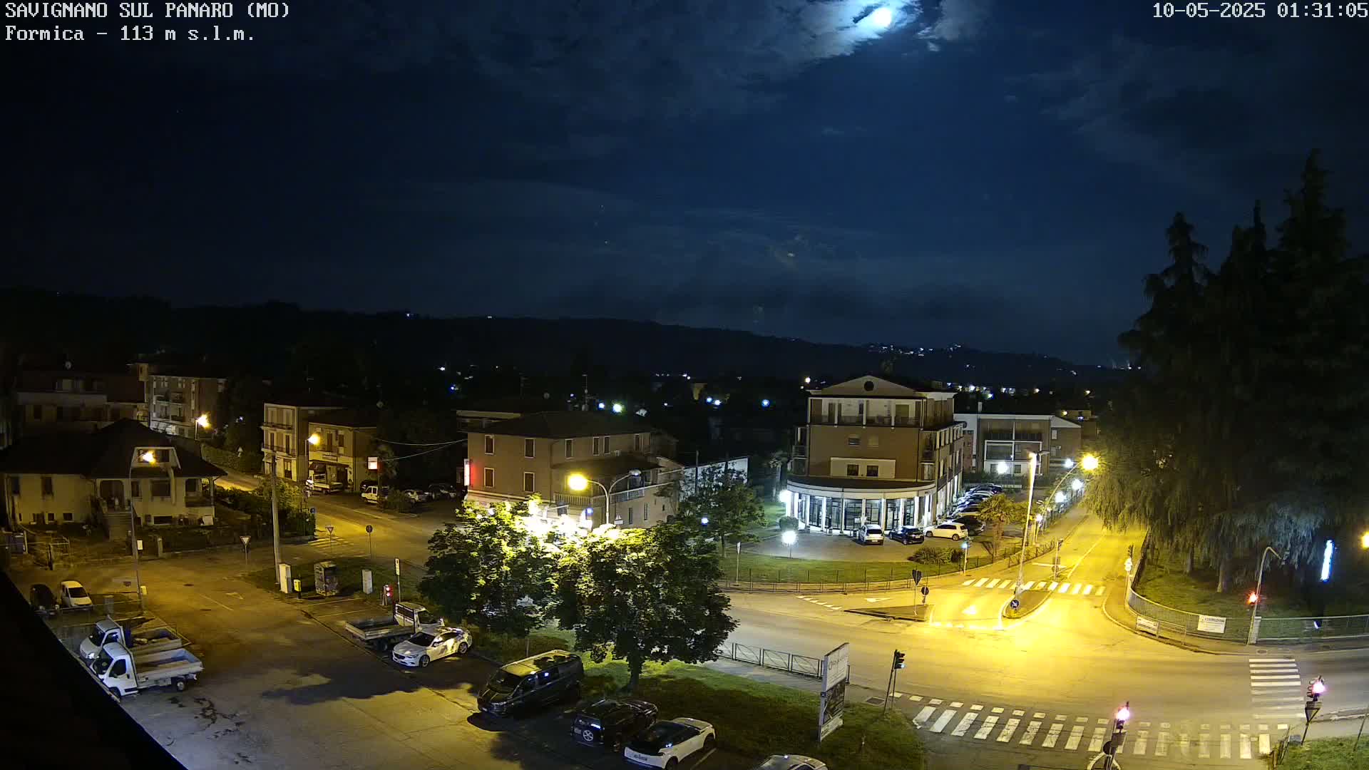A nighttime aerial view of a town square with cars parked and streetlights illuminating the scene under a partly cloudy moonlit sky.