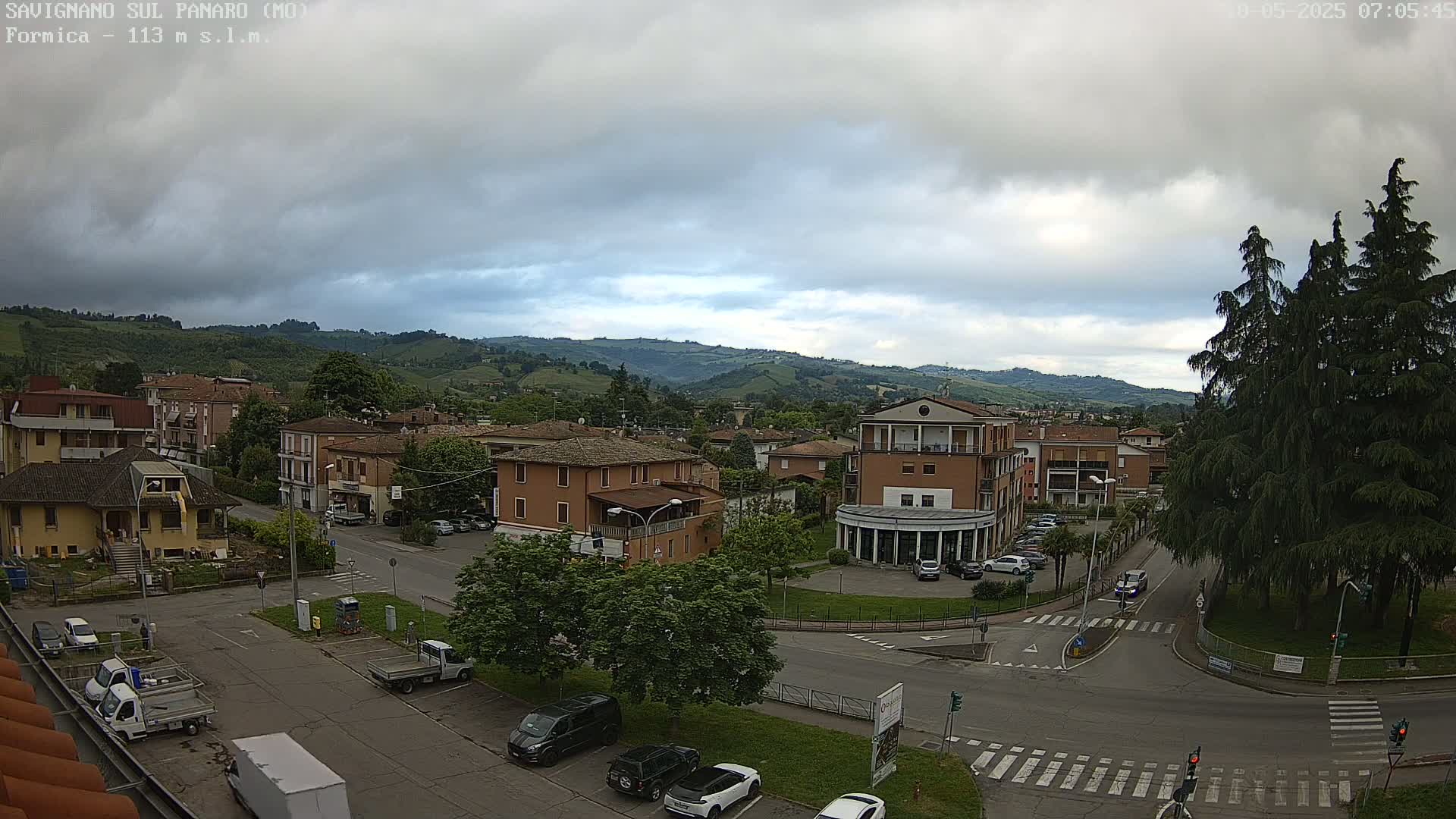 A mostly cloudy day reveals a small town nestled in a valley, surrounded by rolling green hills.