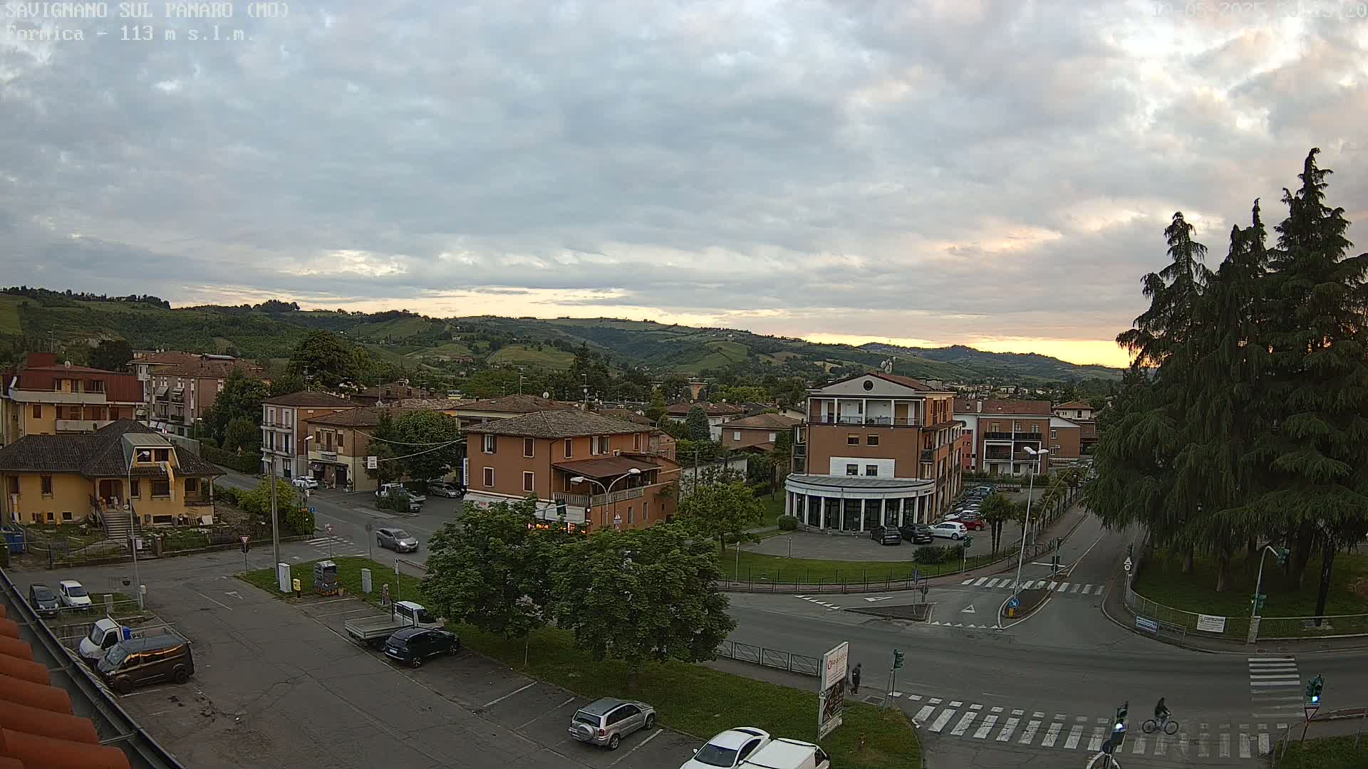A high-angle view of a town square with several buildings, cars parked in lots and on the streets, and green hills in the background under a partly cloudy sky with a hint of sunset.