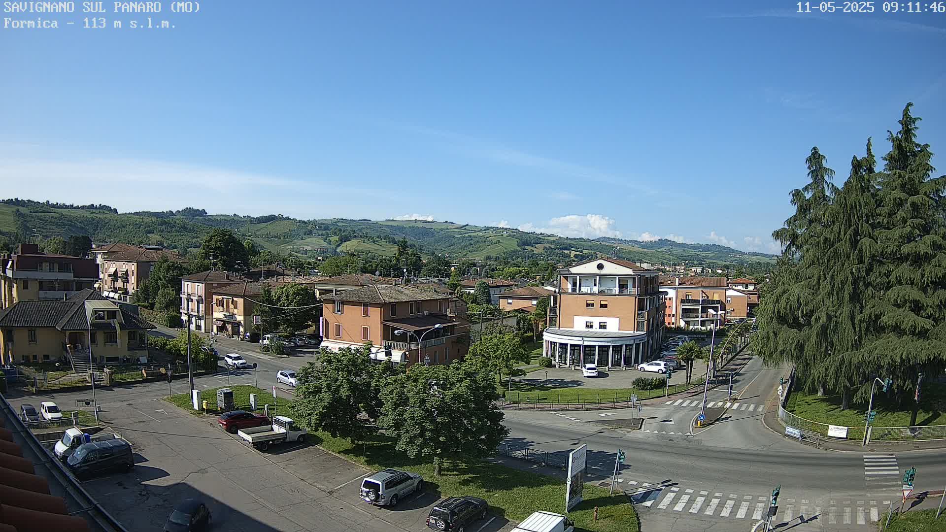 A sunny day reveals a town nestled in a valley, with cars parked along streets and buildings surrounding a small roundabout.