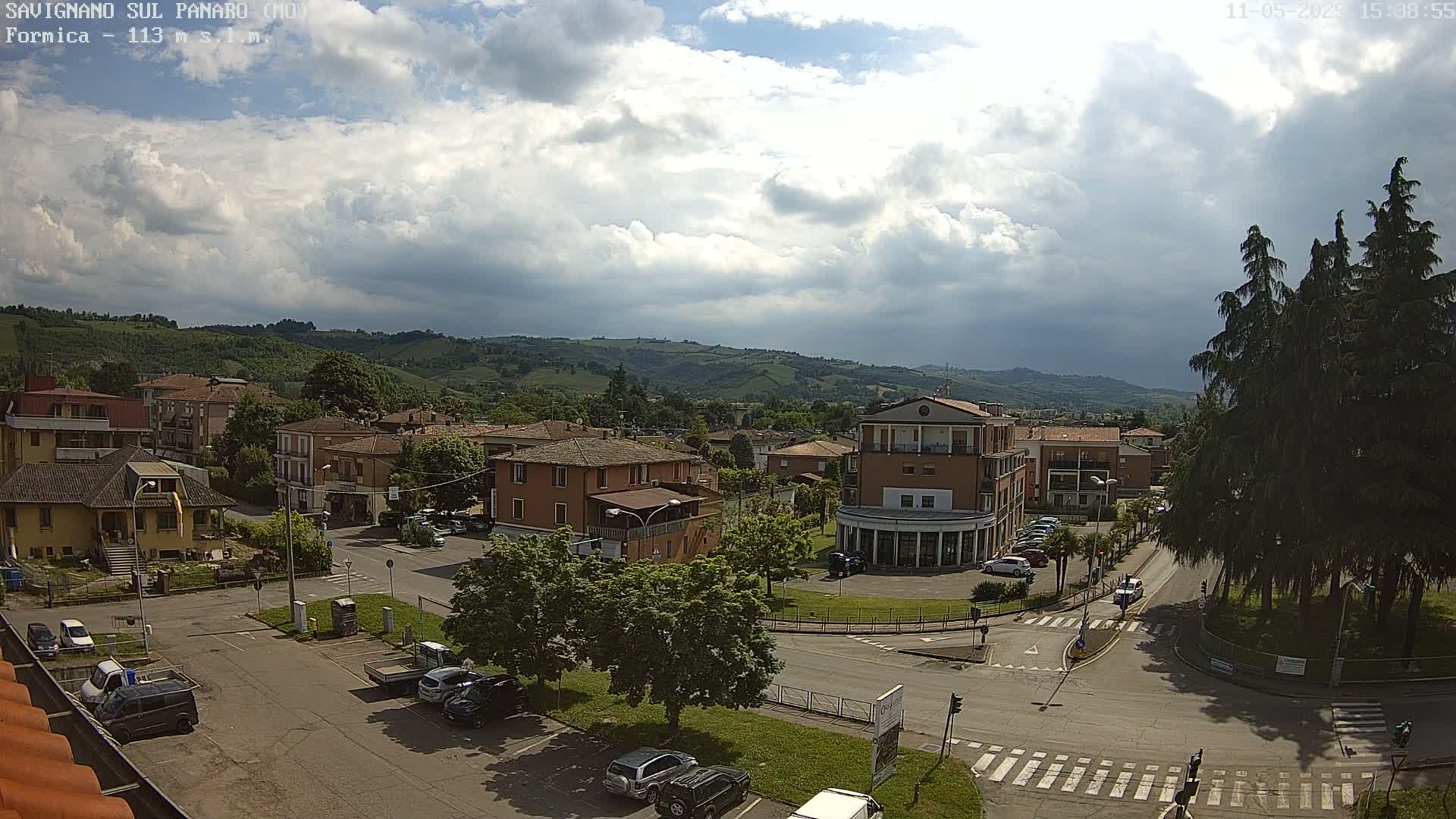 A partly cloudy day overlooks a town nestled in a valley, with cars parked along roadways and trees surrounding a grassy area.