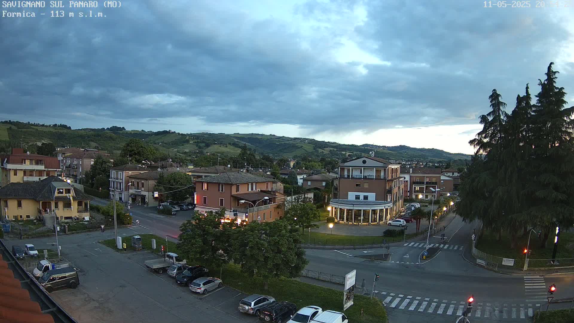 A partly cloudy evening scene overlooks a town square with buildings, cars, and trees, nestled in a valley against a backdrop of rolling hills.