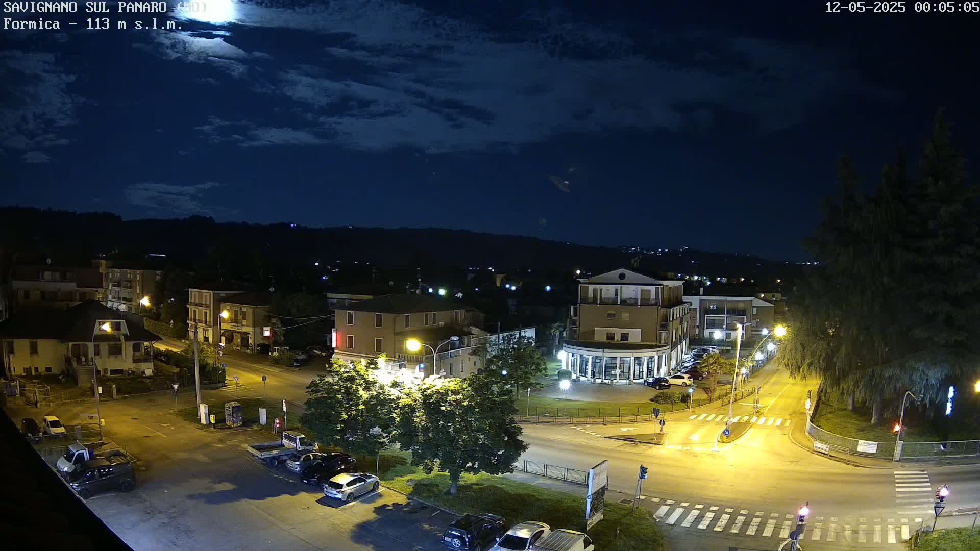 A nighttime, overhead view of a town square with cars parked along the streets, illuminated by streetlights under a partly cloudy sky.