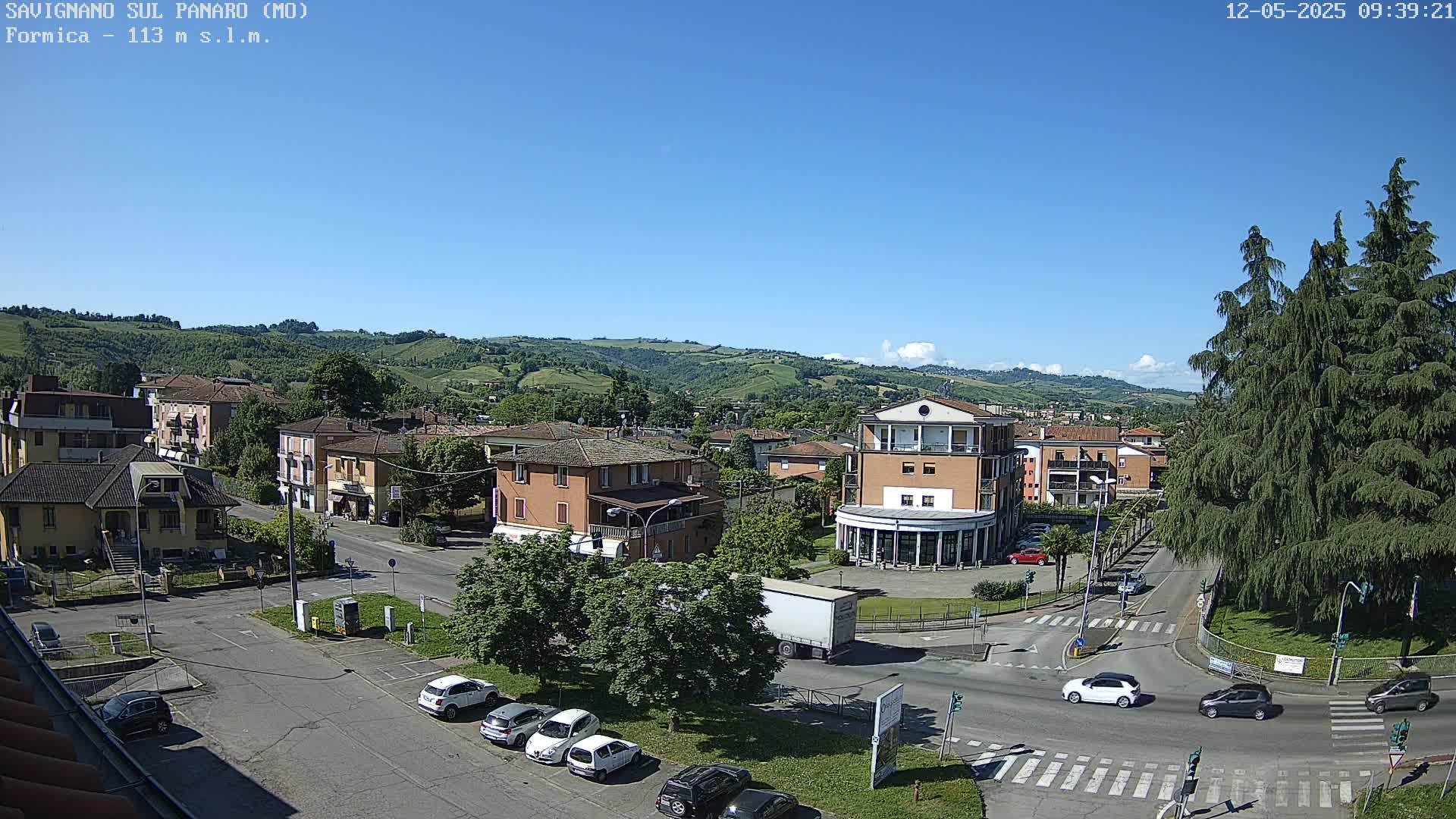 A sunny day reveals a town nestled in a valley, with cars parked along the streets and a few vehicles traversing a crossroads near a large evergreen tree.