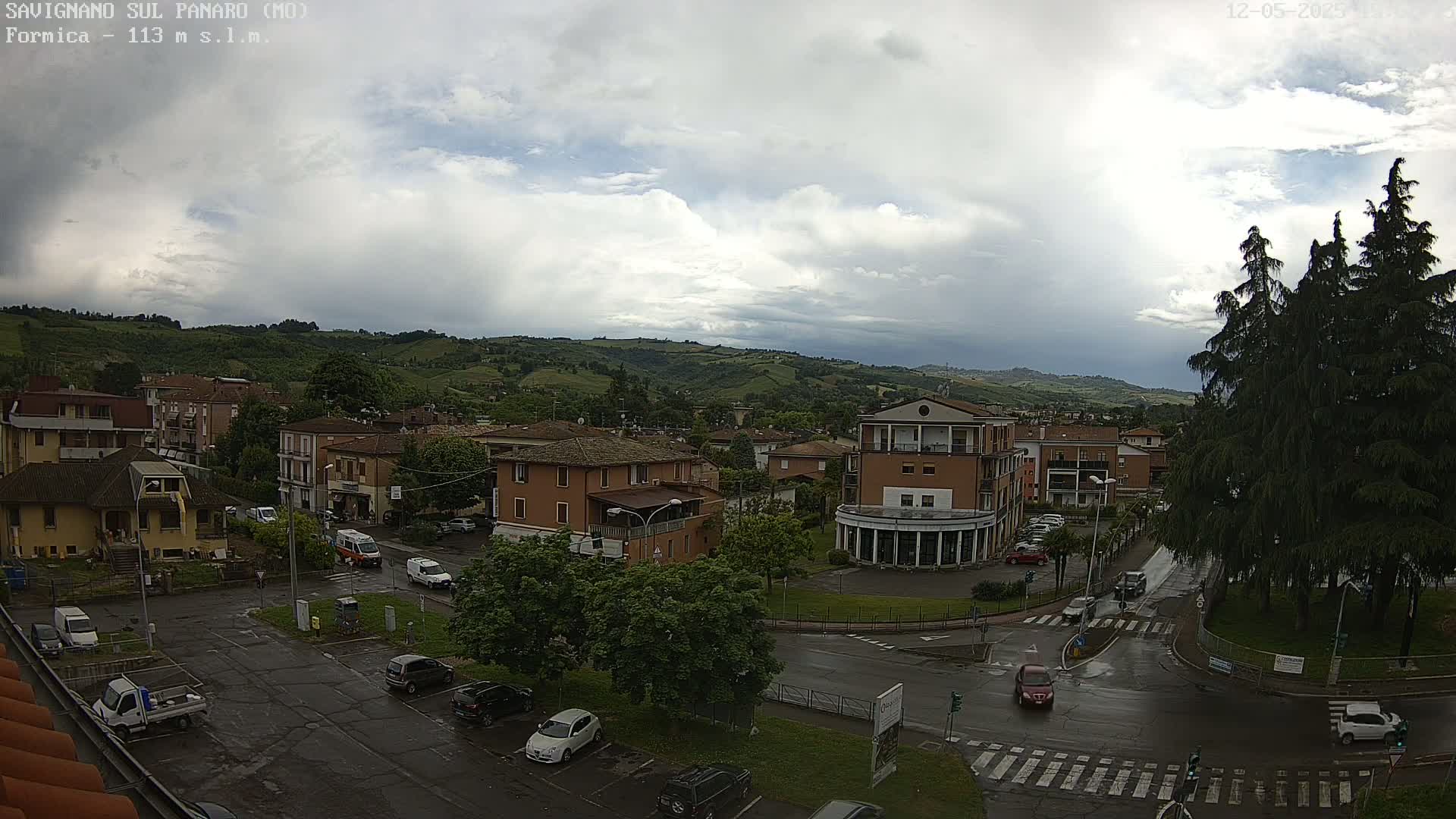A wet town square with several cars and buildings is nestled in a valley under a cloudy sky.