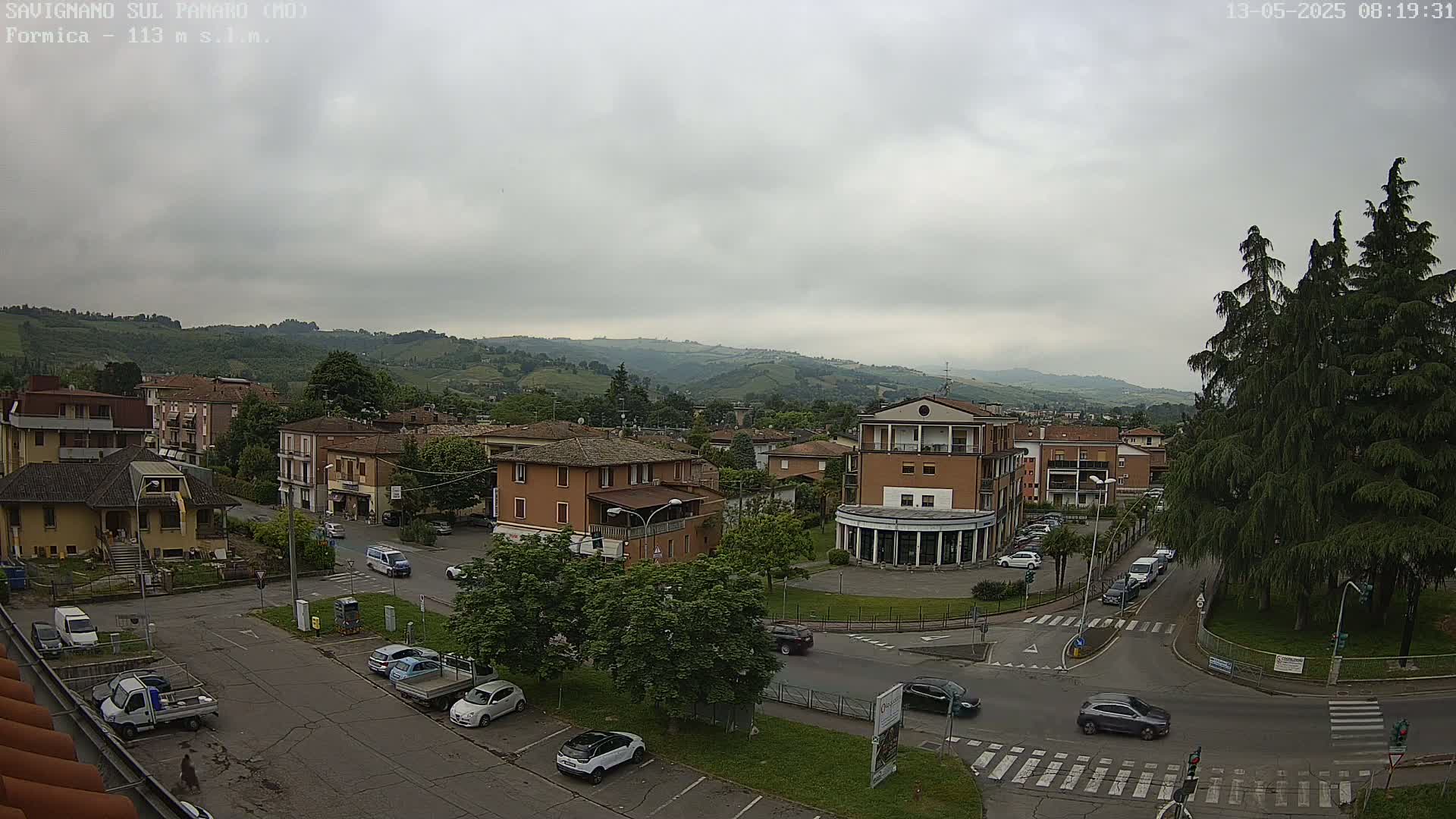 A mostly cloudy day reveals a town square with several cars parked and driving, surrounded by low buildings and lush green hills in the background.
