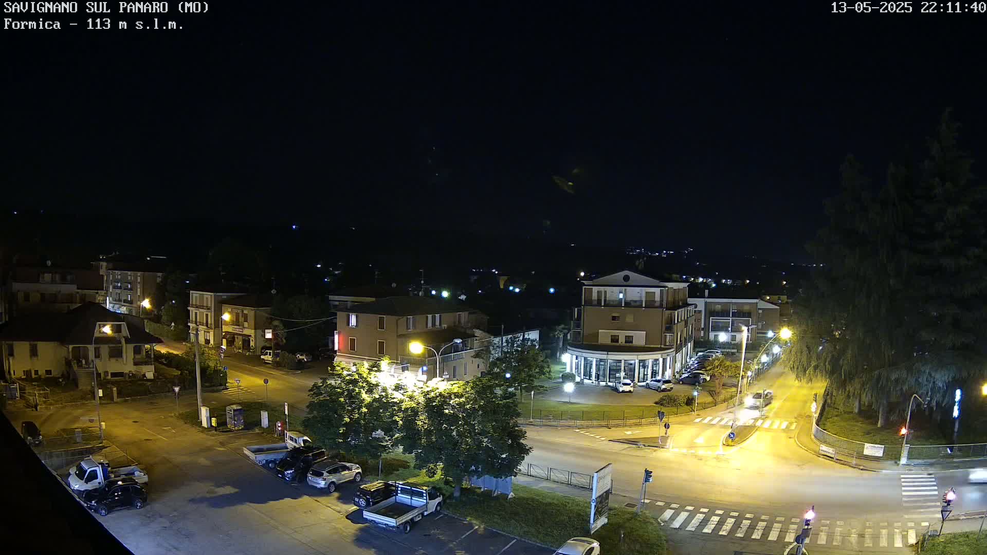 A nighttime aerial view of a town square with cars parked and driving on roads, illuminated by streetlights under a clear, dark sky.