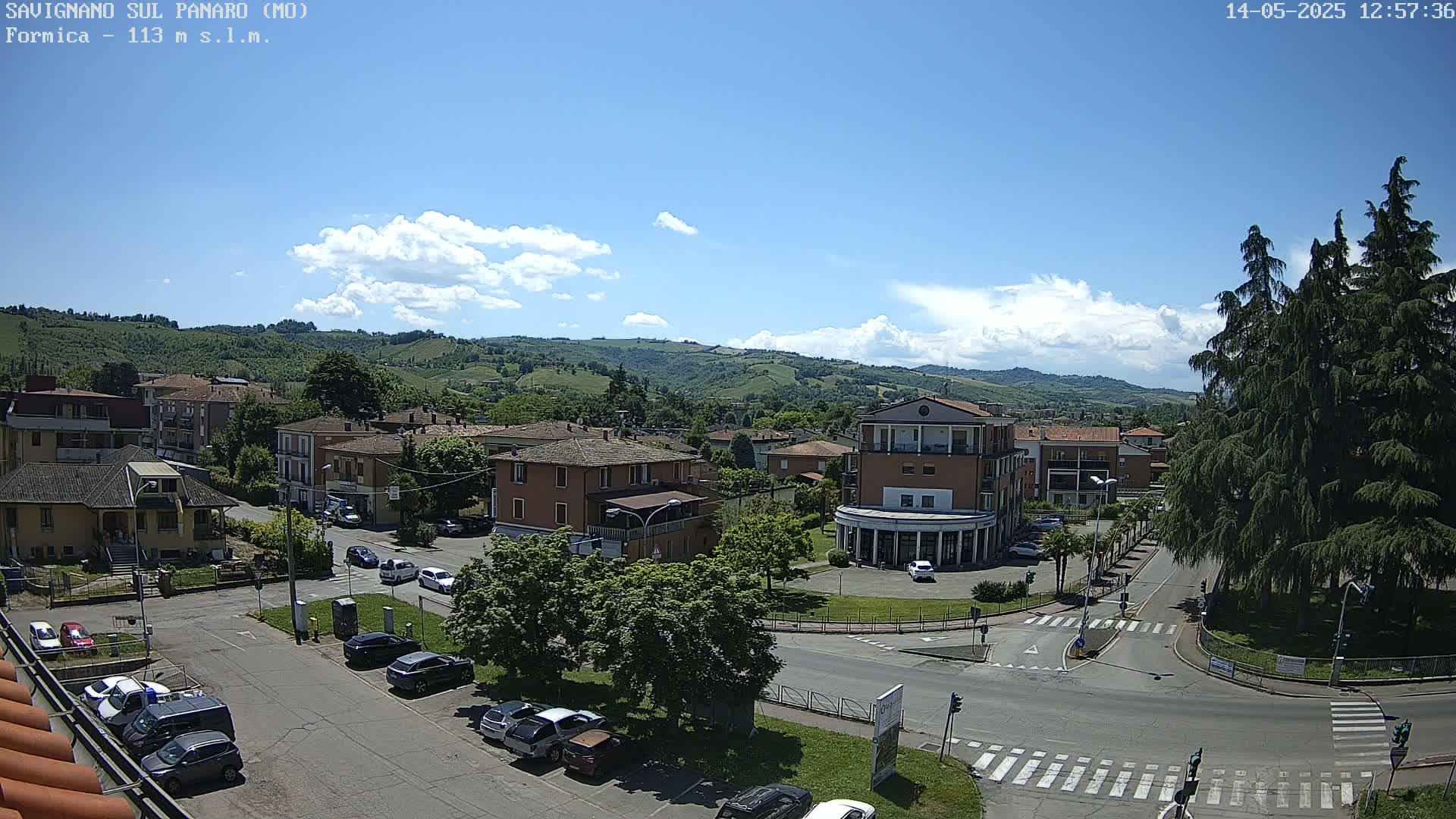 A mostly sunny day reveals a small town nestled in a valley, with cars parked along streets and buildings surrounded by greenery.