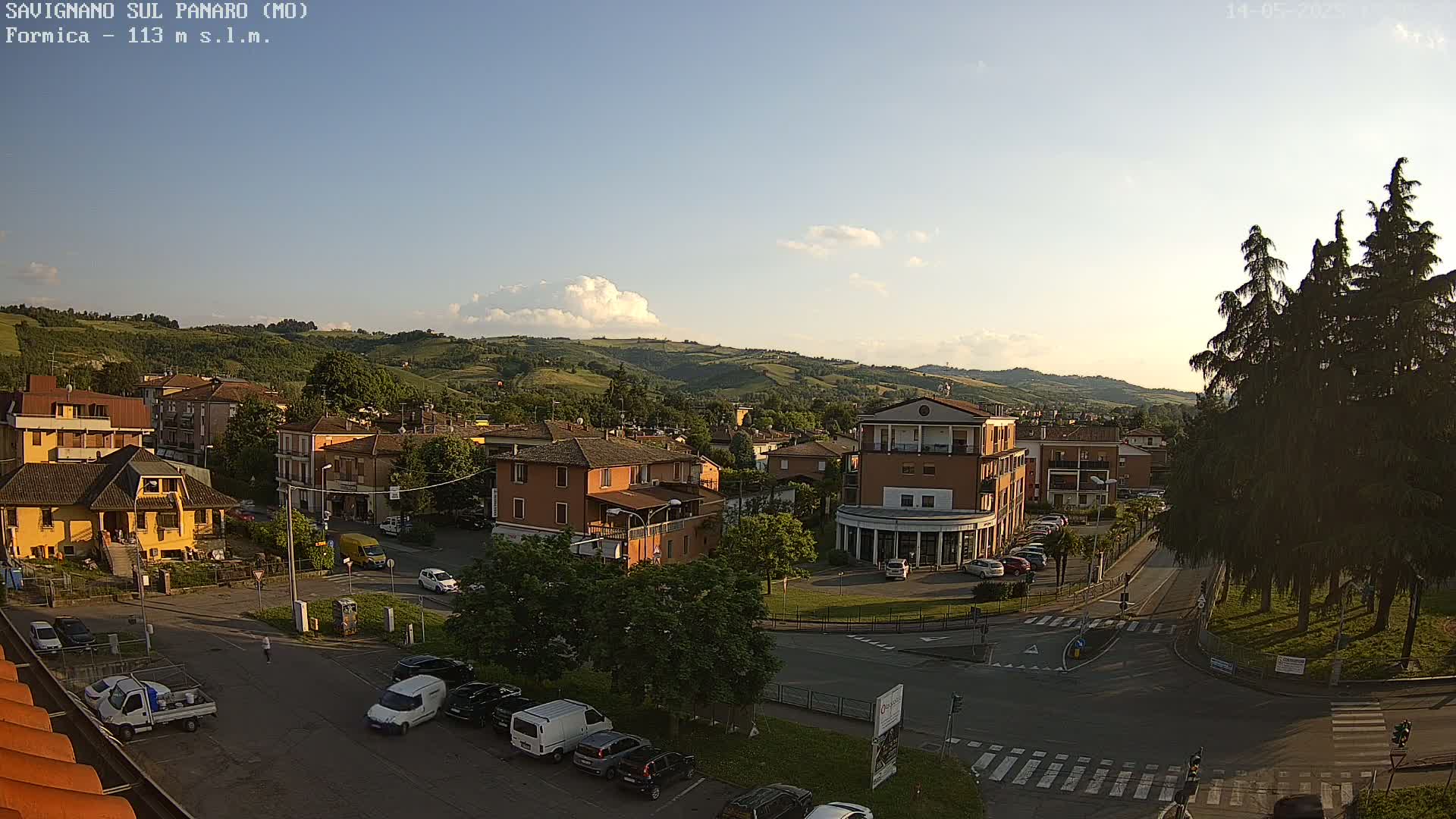 A partly sunny view of a small town nestled in a valley, with cars parked along the streets and buildings in muted earth tones.