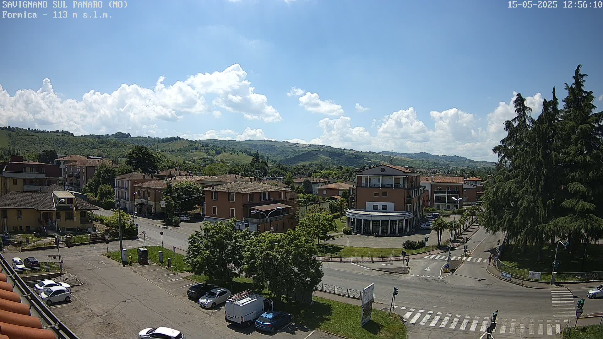 A partly sunny day reveals a town nestled in a valley, surrounded by rolling hills and green vegetation, with cars parked in a street-level lot and several buildings visible.