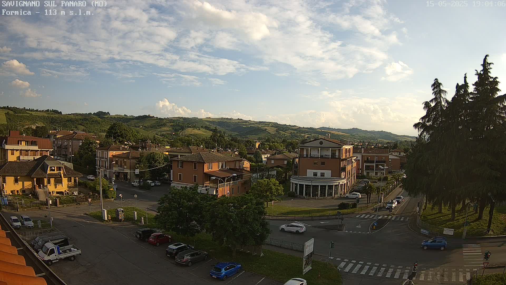 A partly sunny view of a small town nestled in a valley, with cars parked and driving on roads that intersect at a roundabout.