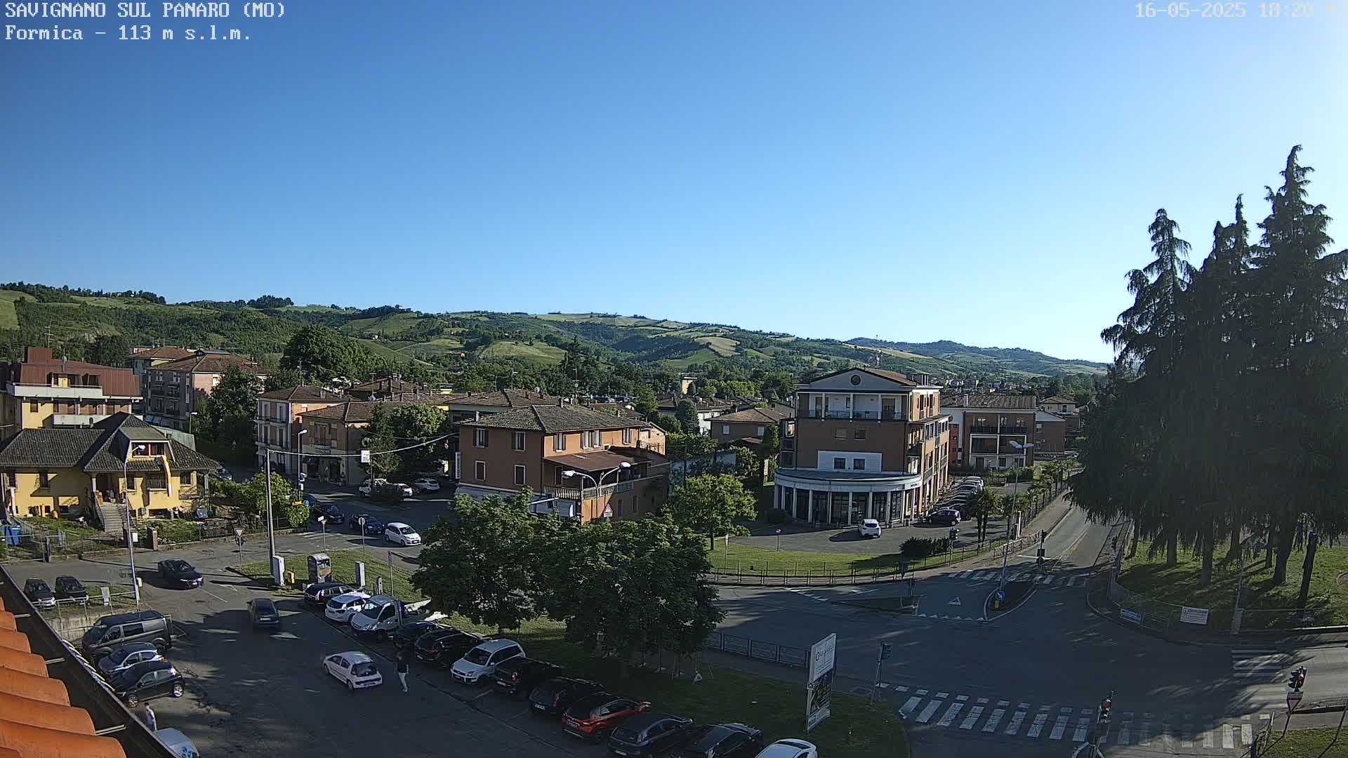 A sunny day reveals a town nestled in a valley, with buildings, cars parked in lots, and green hills in the background.