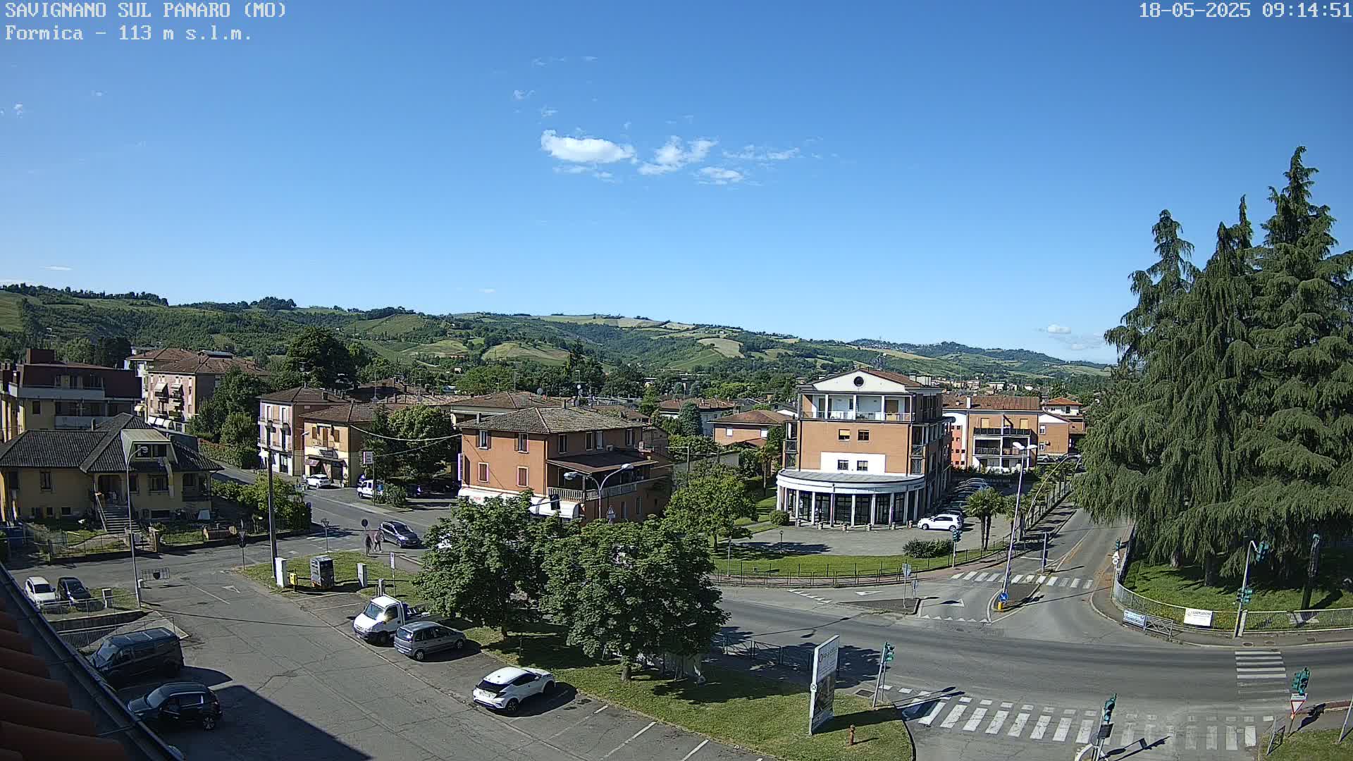 A sunny day overlooks a town square with cars parked and a few pedestrians, nestled in a valley surrounded by rolling green hills.