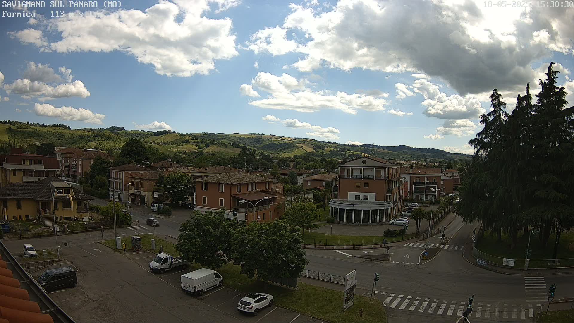 A partly cloudy day reveals a view of a town nestled in a valley, with cars parked in a lot and roads intersecting in the foreground.