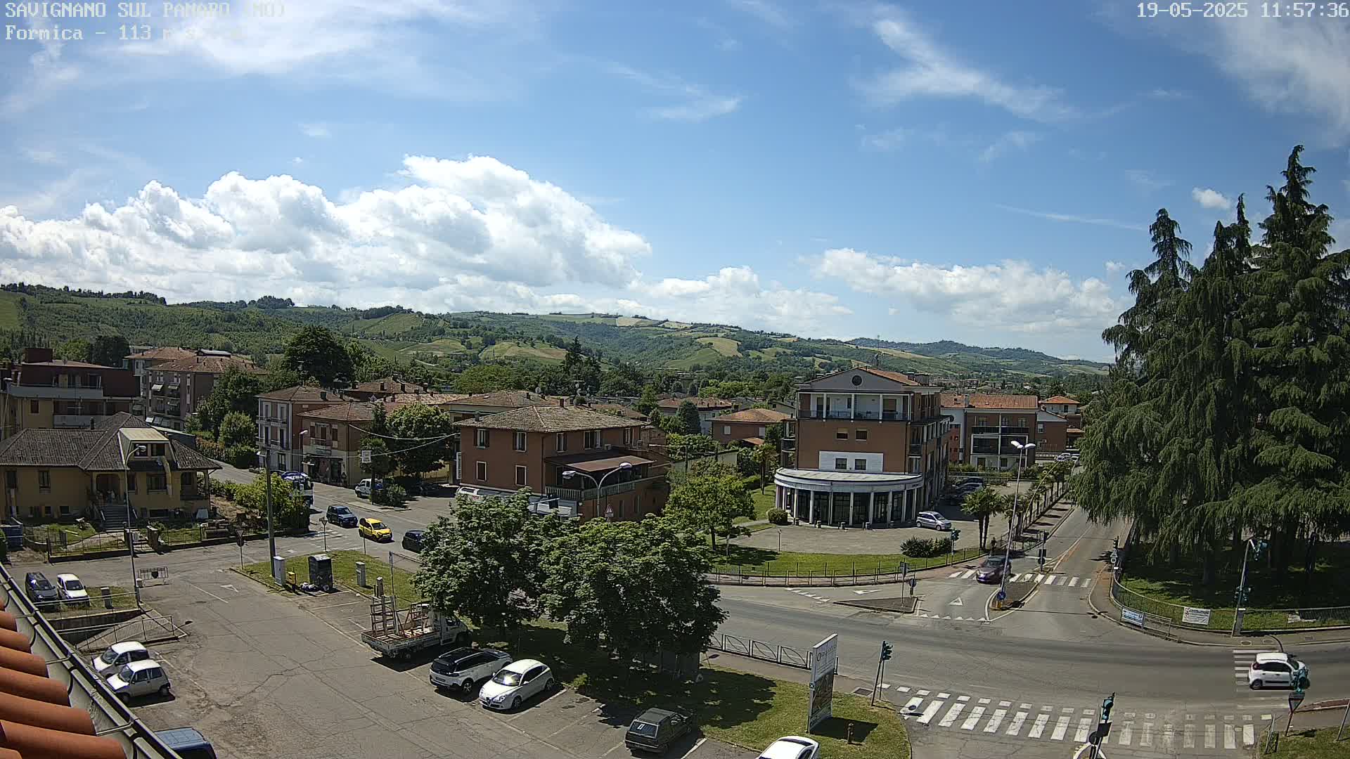 A sunny day with partly cloudy skies overlooks a town square with buildings, cars, and trees, set against a backdrop of rolling green hills.