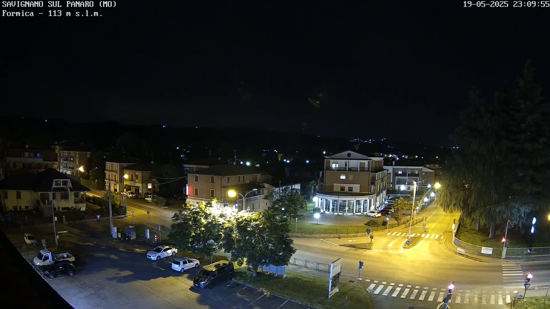 A nighttime, high-angle view of a town square with cars parked and a few cars driving, surrounded by buildings and trees under a clear, dark sky.