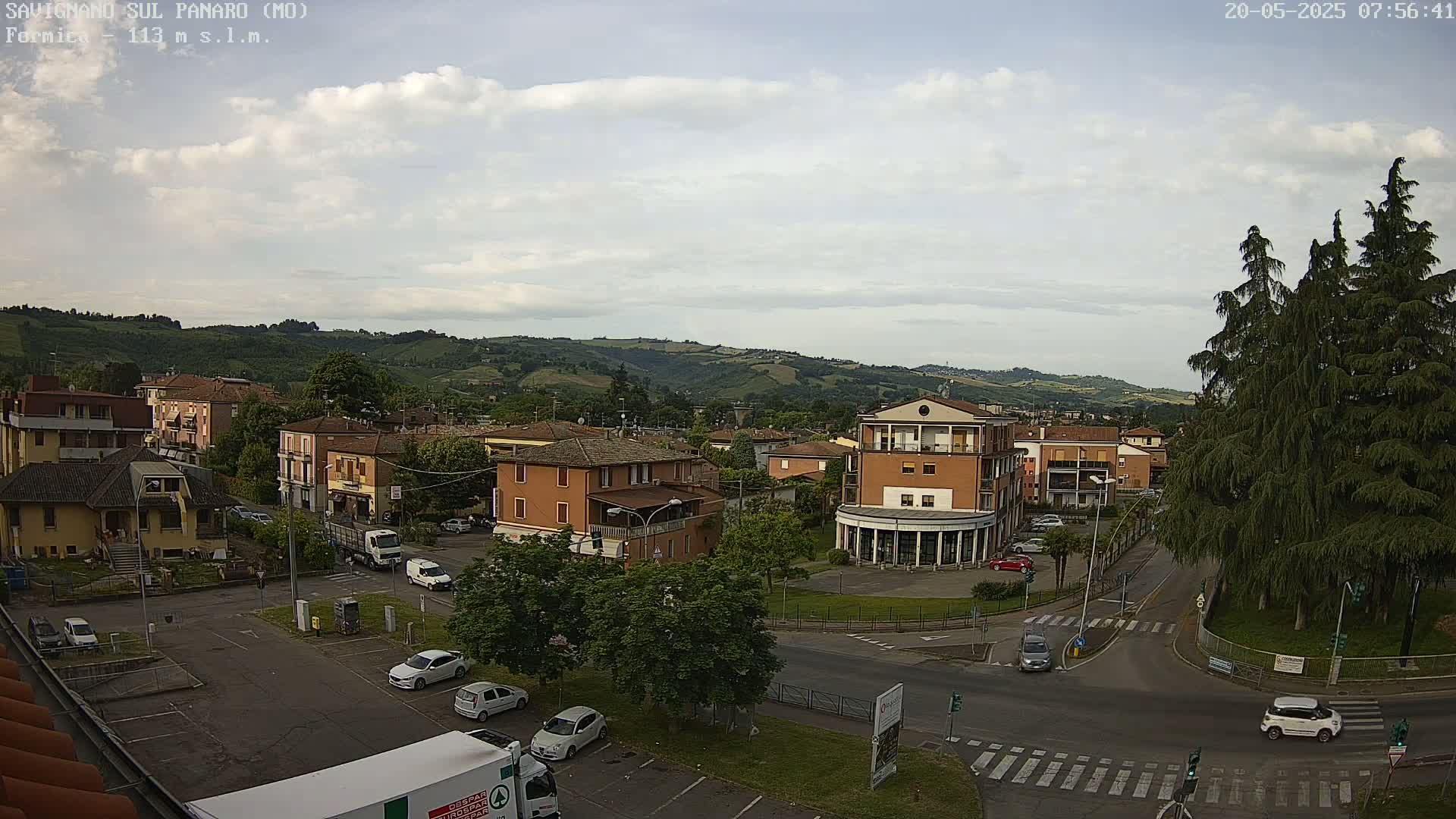 A partly cloudy day reveals a small town nestled in a valley, surrounded by rolling hills, with cars parked in a nearby lot.