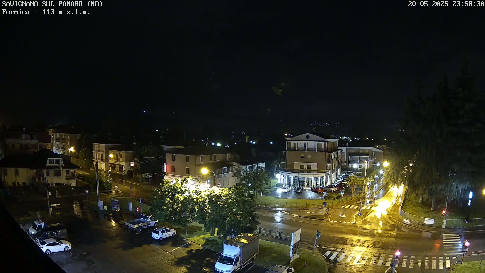 A nighttime, rain-slicked town square with several buildings and parked cars is illuminated by streetlights.