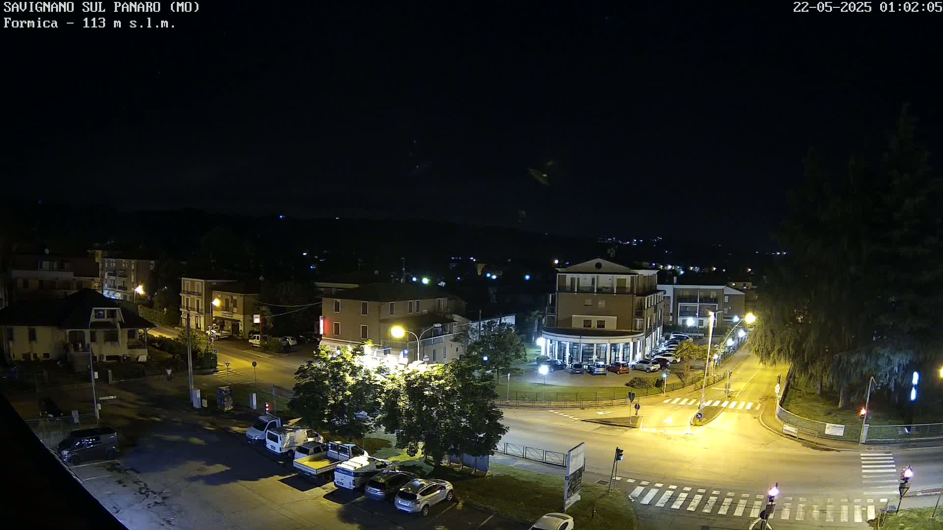 A nighttime, high-angle view of a town square with cars parked around its perimeter and several buildings lit by streetlights.