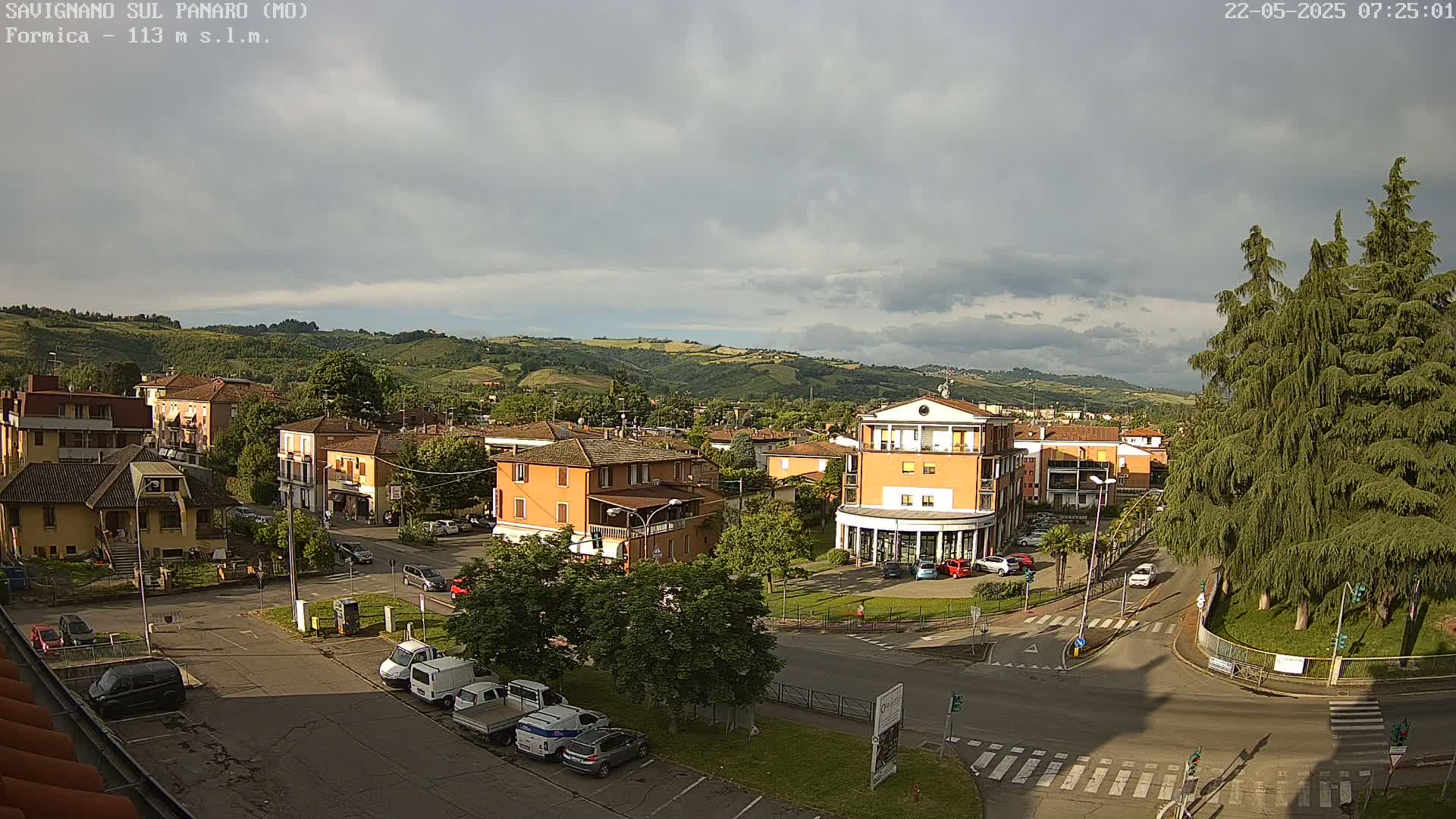 An aerial view of a town square with buildings, parked cars, and green hills in the background under a partly cloudy sky.